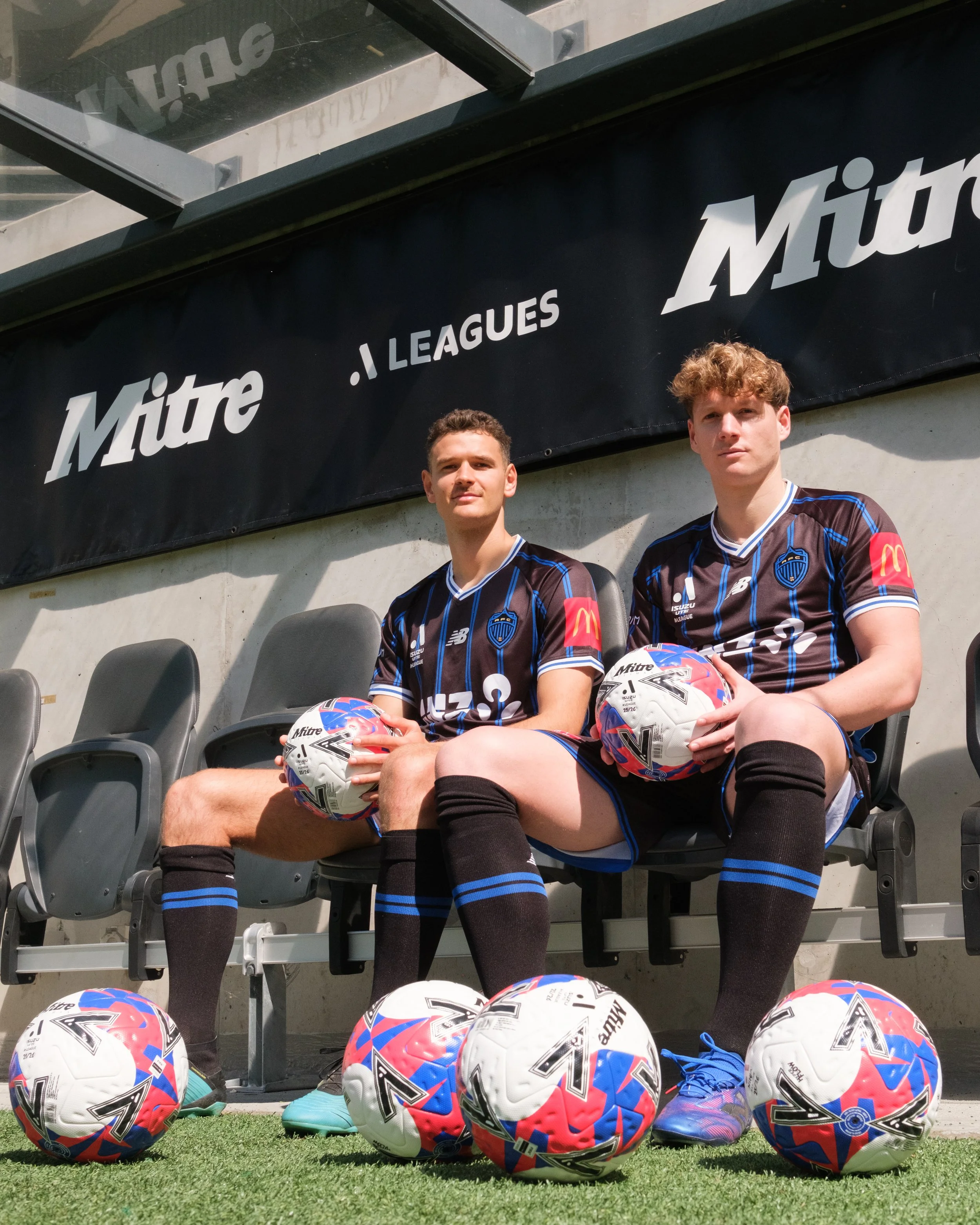 Two soccer players in black and blue uniforms sitting on stadium seats, holding soccer balls, with several more soccer balls on green turf in the foreground, and a large black banner with white text behind them.