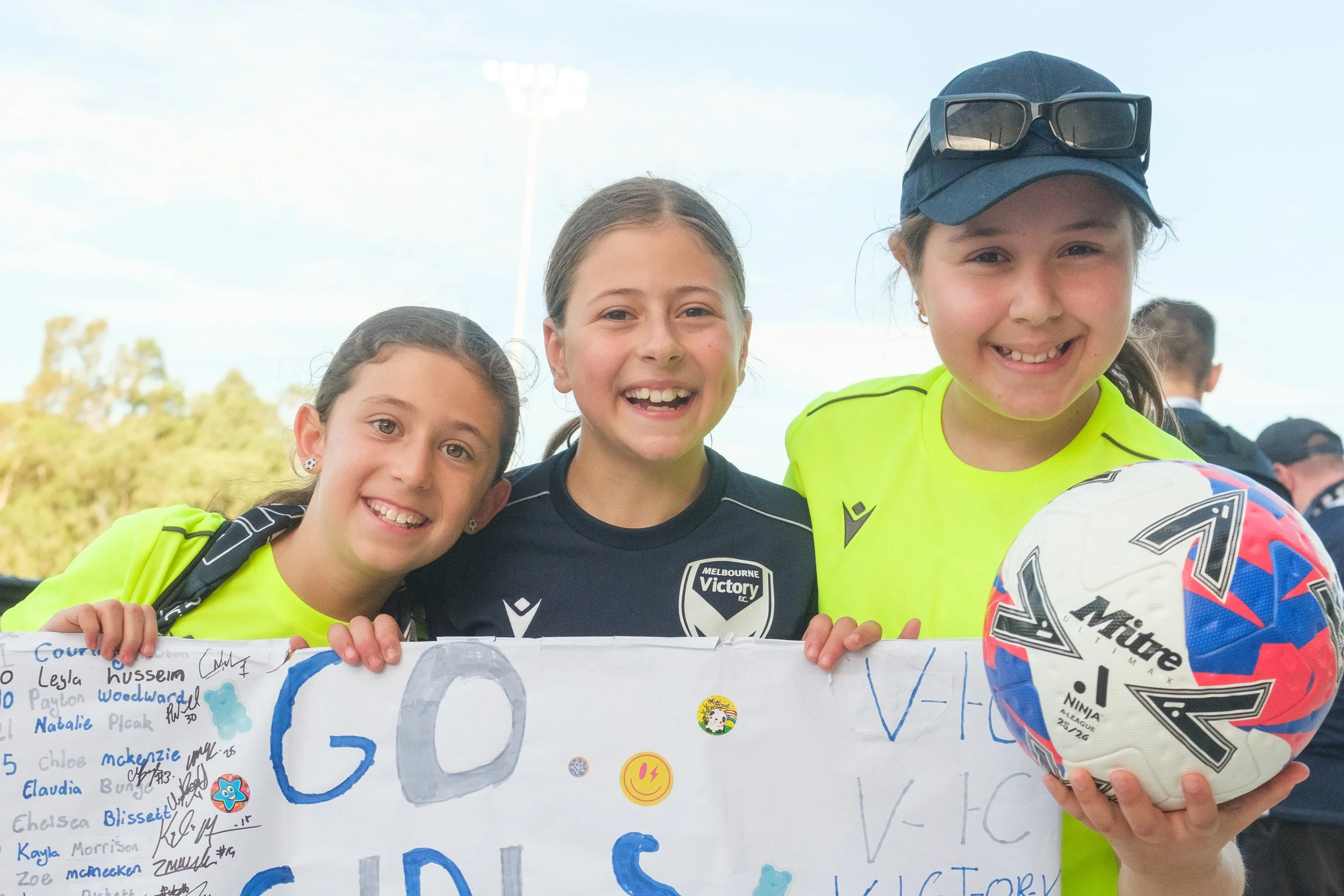 Three young girls smiling at a soccer event, holding a colorful poster with signatures and drawings, with blue sky and trees in the background.