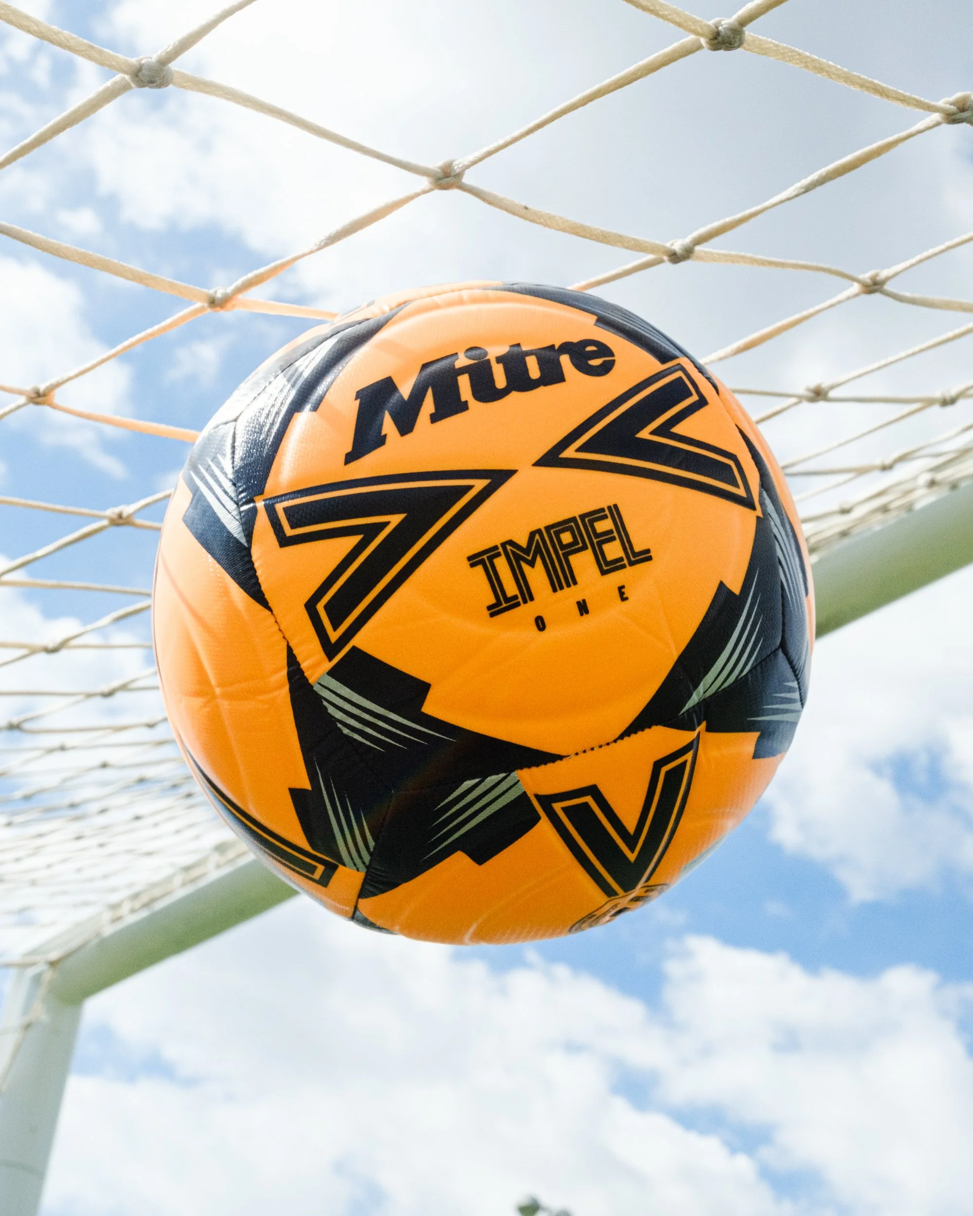 Close-up of an orange and black sports ball with the brand name 'Müte' and the words 'Tampel One,' hanging on a soccer goal net against a cloudy sky.