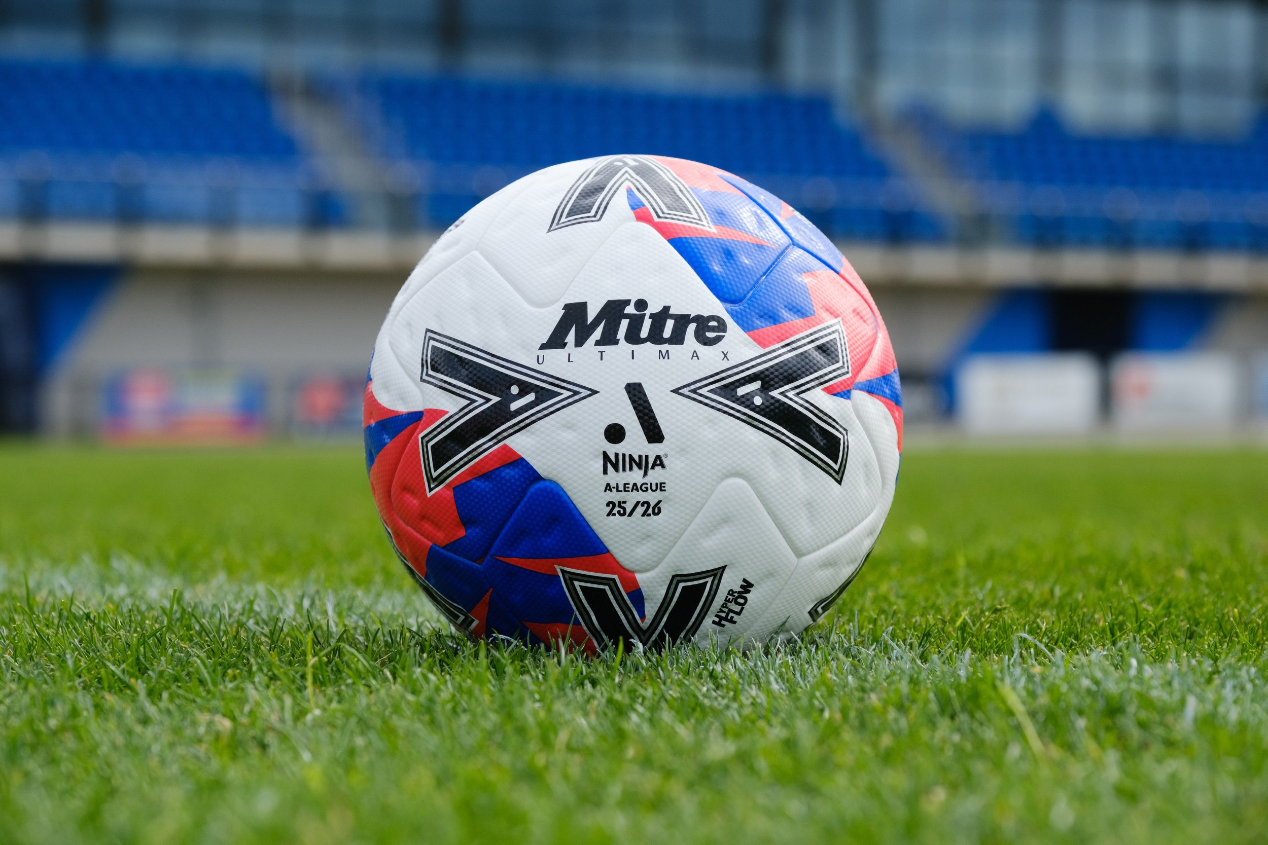 A white, red, and blue soccer ball on a grassy field. The ball has various branding and logo markings, including 'Mitre,' 'Ninja,' and 'A-League,' with a background of stadium seating.