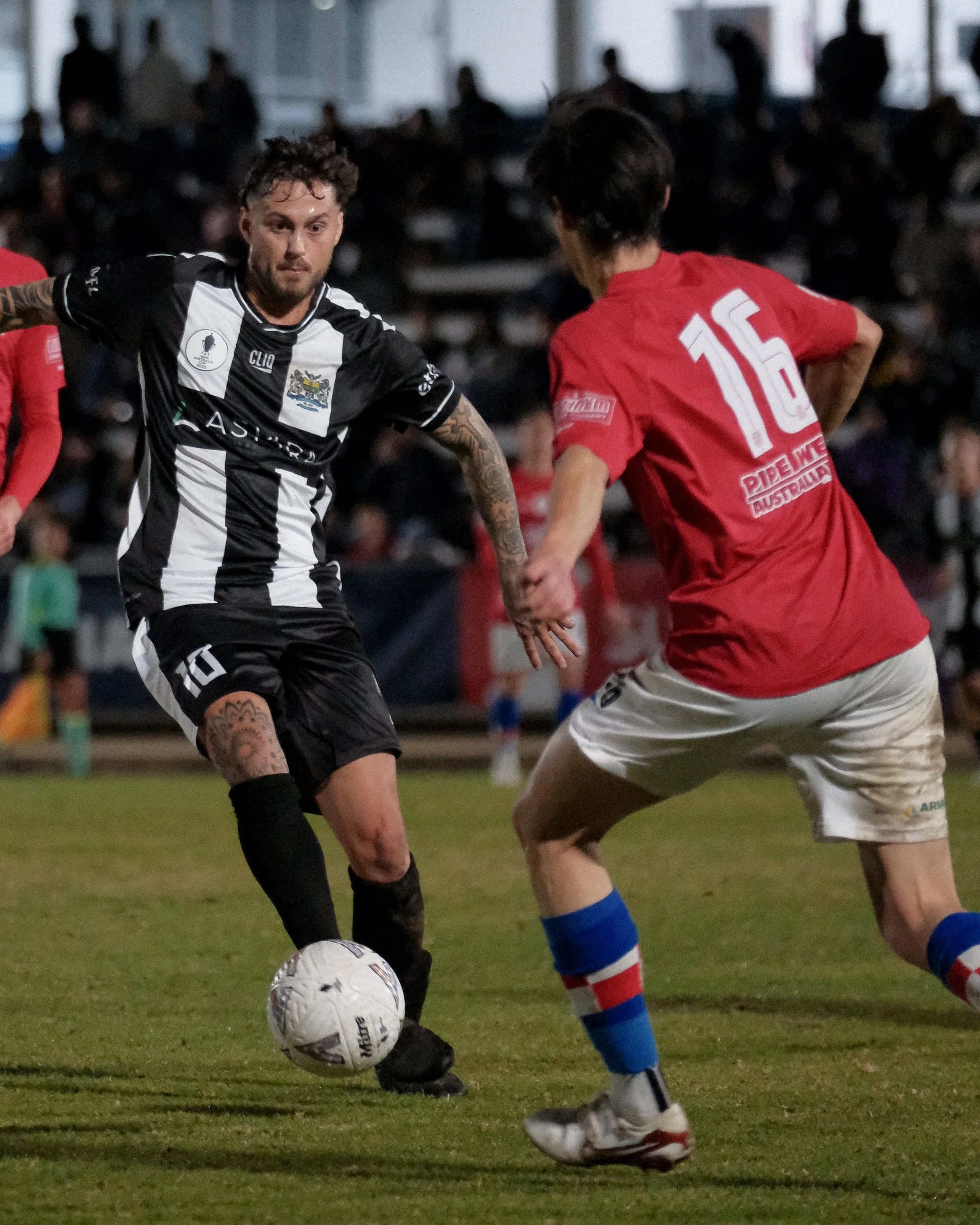 Two soccer players are battling for control of the ball on the field. One player, in a black and white jersey with tattoos on his arm, is kicking the ball. The other player, in a red jersey with the number 15, is approaching. The background shows a c