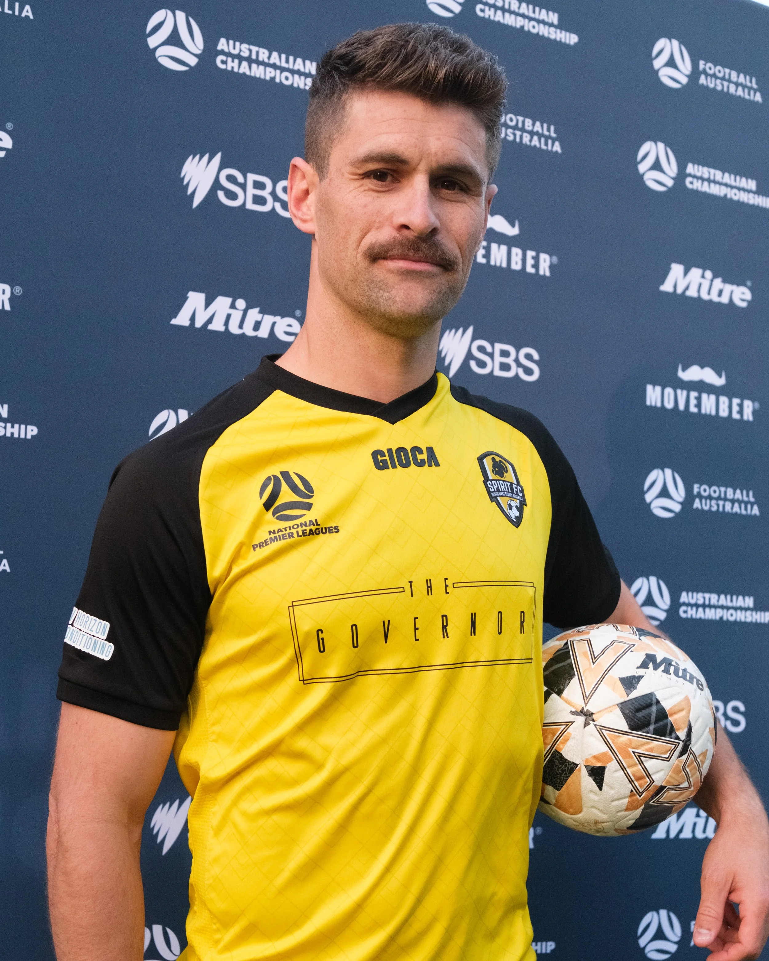 A man in a yellow and black sports jersey stands in front of a blue backdrop with white text and logos, holding a patterned sports ball under his arm.