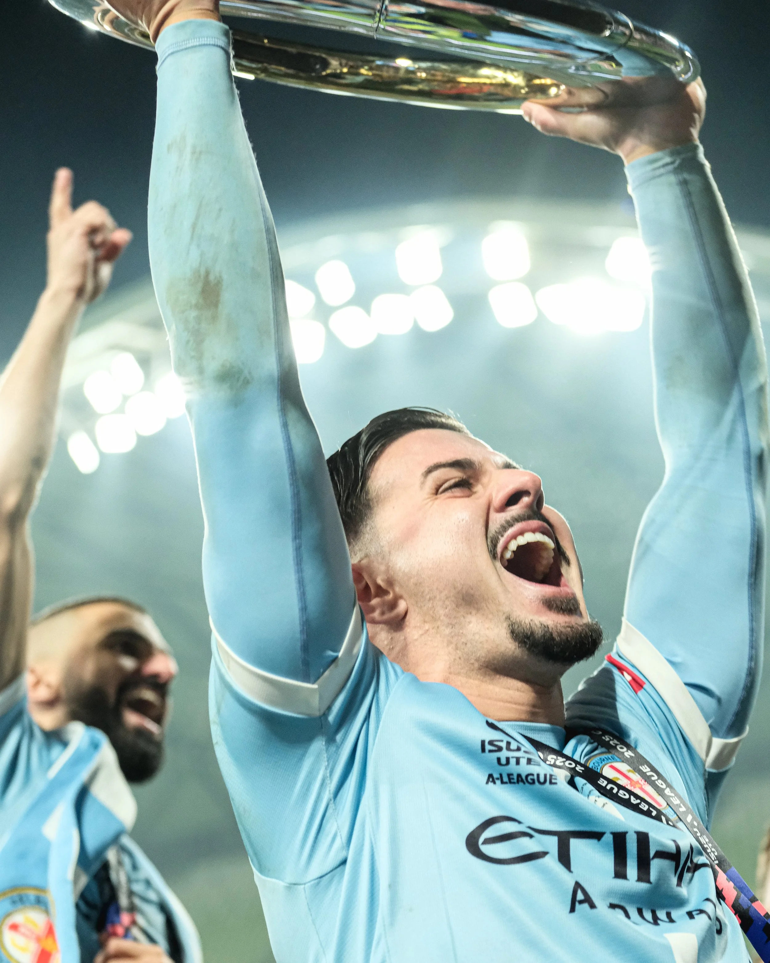 A man in a light blue sports jersey is holding a silver trophy above his head, celebrating with his eyes closed and mouth open in joy. Other people in similar jerseys are visible in the background, also celebrating.