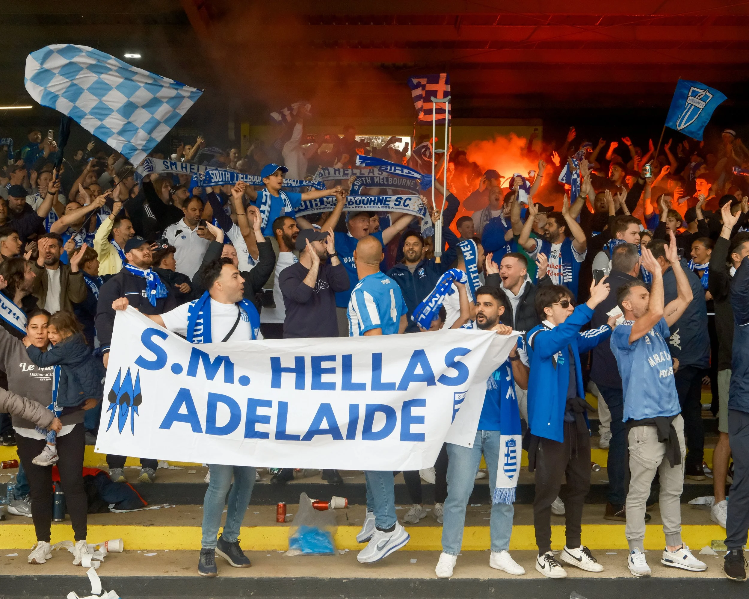 Crowd of fans wearing blue and white, holding scarves and flags, celebrating at a sports event. Smoke and flares in the background.