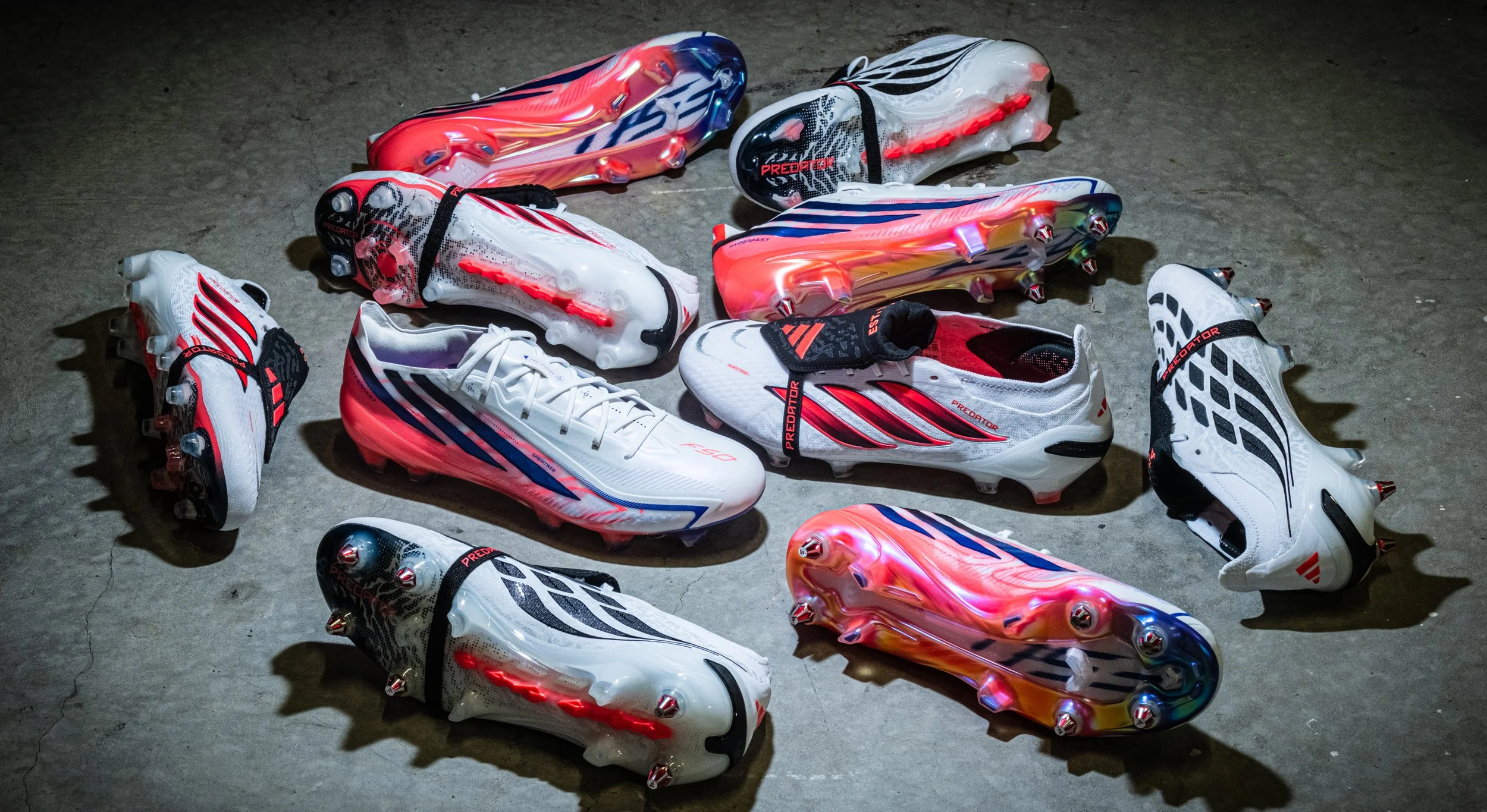 A collection of white and colorful soccer cleats with red and black accents, arranged on a concrete floor. Some cleats have light-up soles.