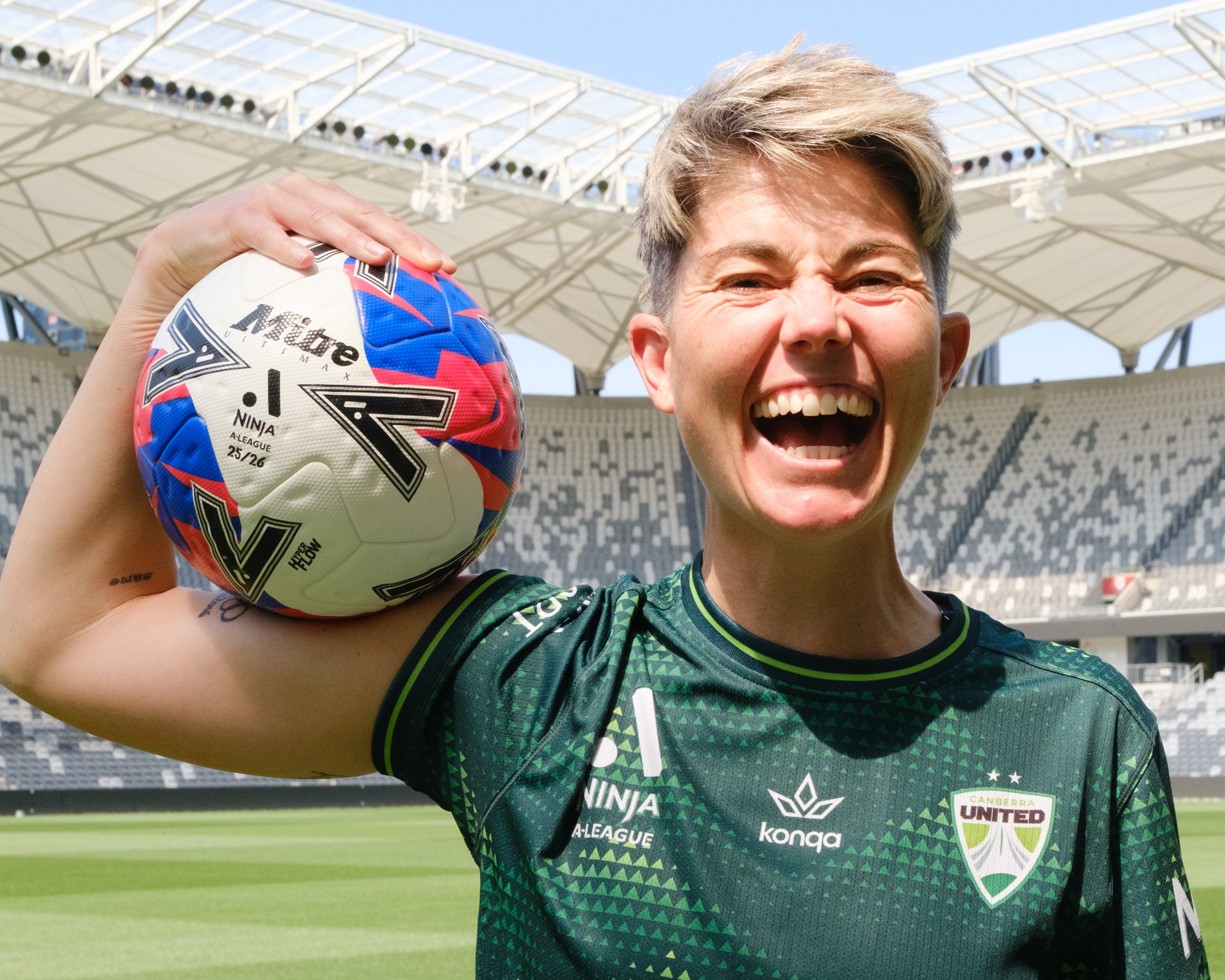 A person in a green sports jersey holding a colorful soccer ball, smiling, with a stadium background.