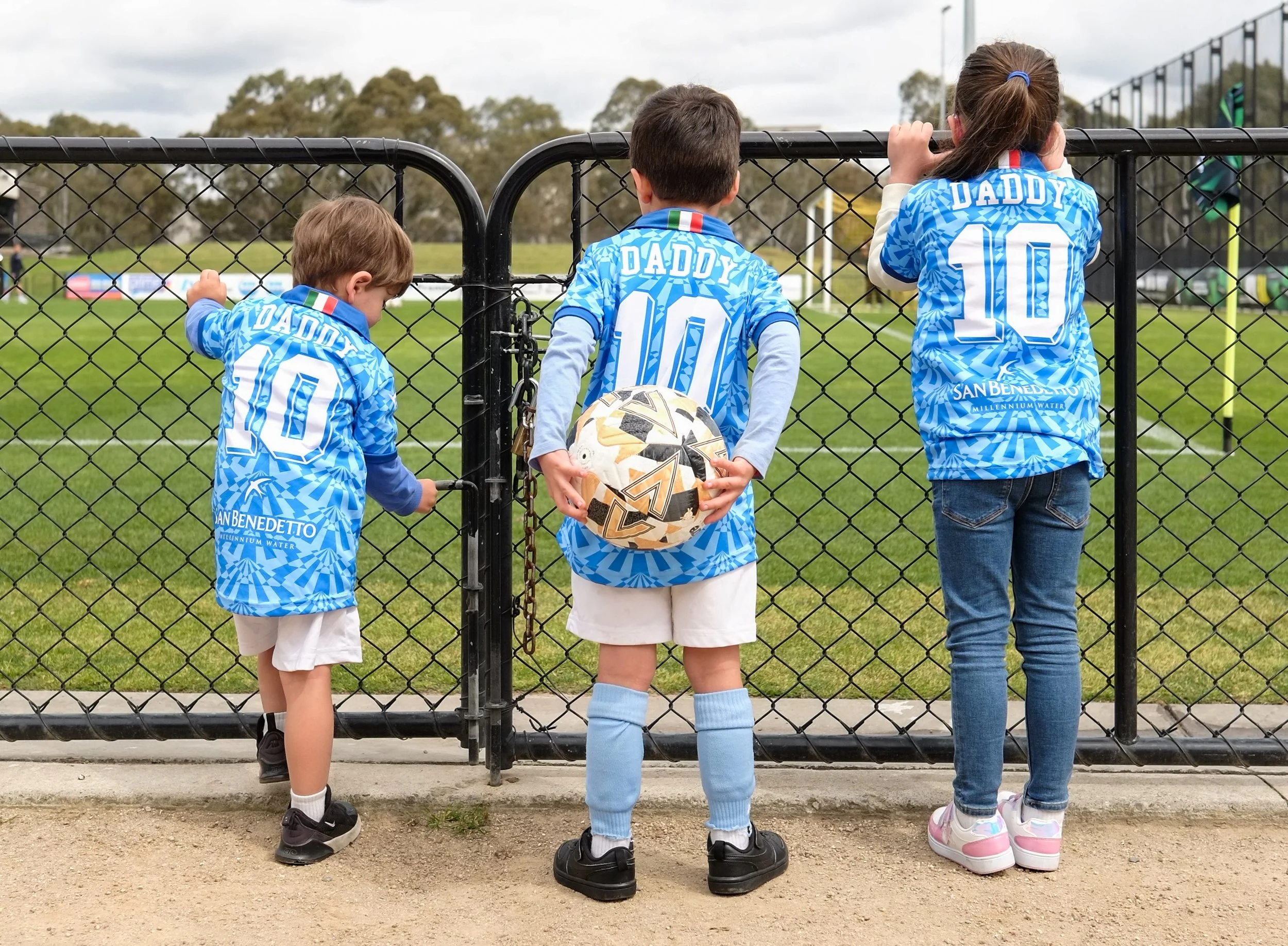 Three children and one adult standing behind a black chain-link fence at a sports field, all wearing matching blue sports jerseys with 'DADDY' and the number 10 on the back, watching a game.