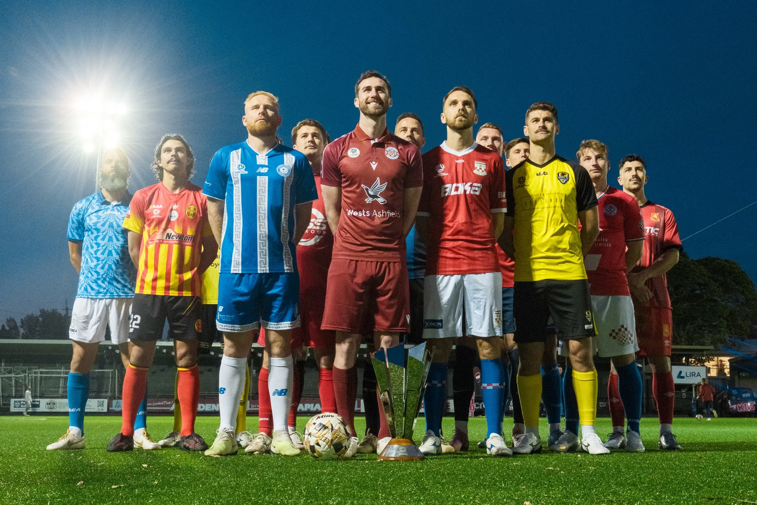 A group of soccer players standing on a field during evening or night, posing for a photo, with a trophy and a soccer ball in front. They are wearing colorful uniform jerseys and shorts with different team logos.
