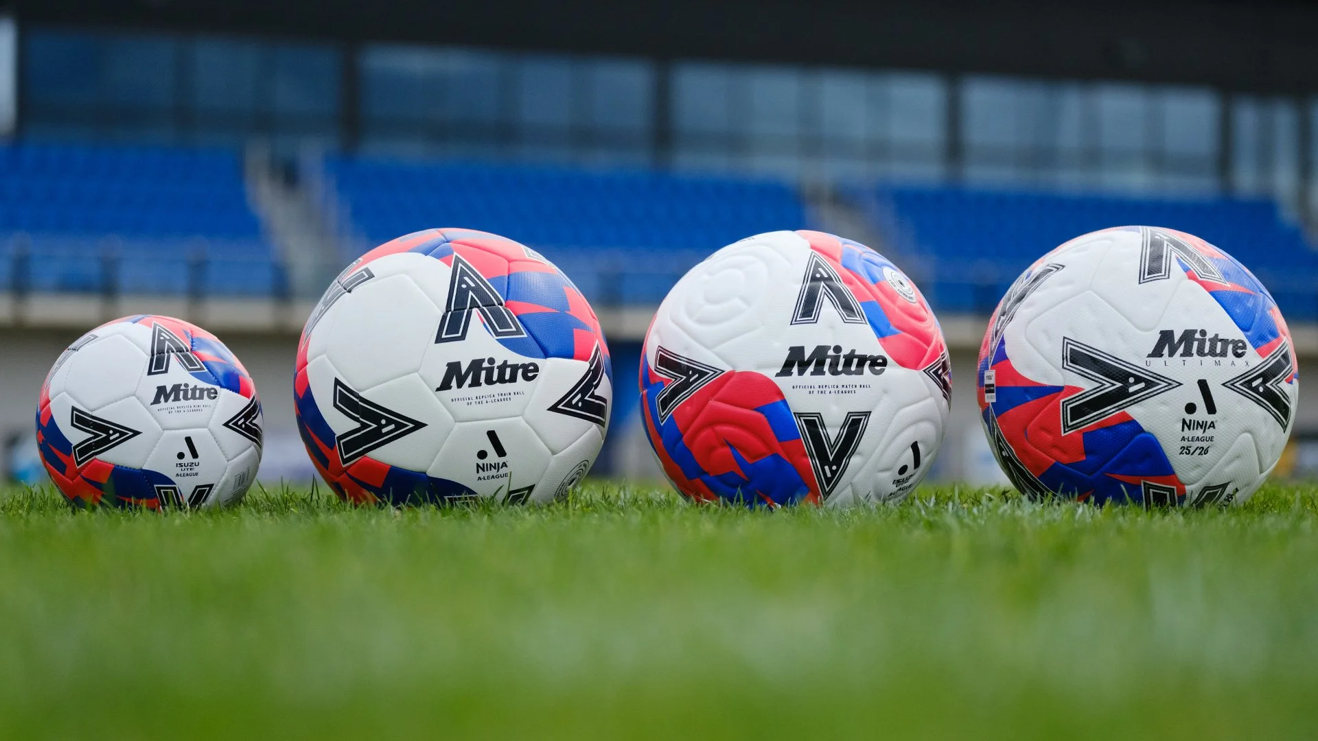 Four soccer balls with a red, white, and blue design on a grass field, with a stadium in the background.