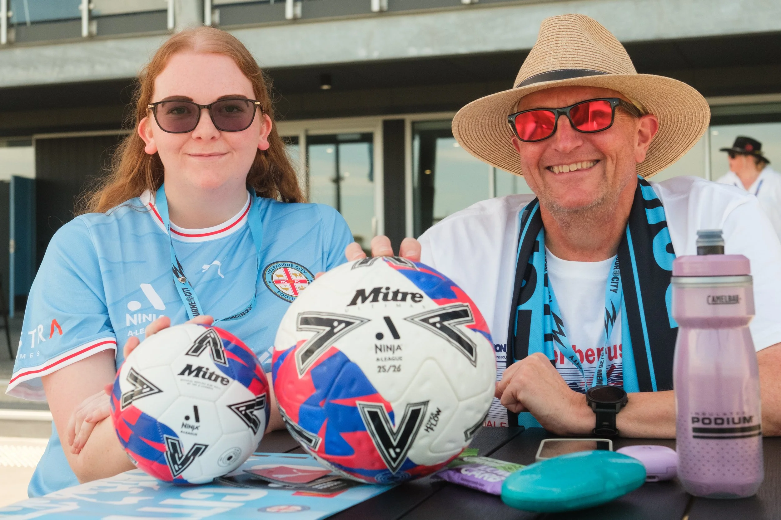 A young woman and an older man sitting at a table, smiling, with sports helmets and water bottles on the table. The young woman is wearing a light blue sports jersey and glasses, and the older man is wearing a straw hat, sunglasses, and a white shirt
