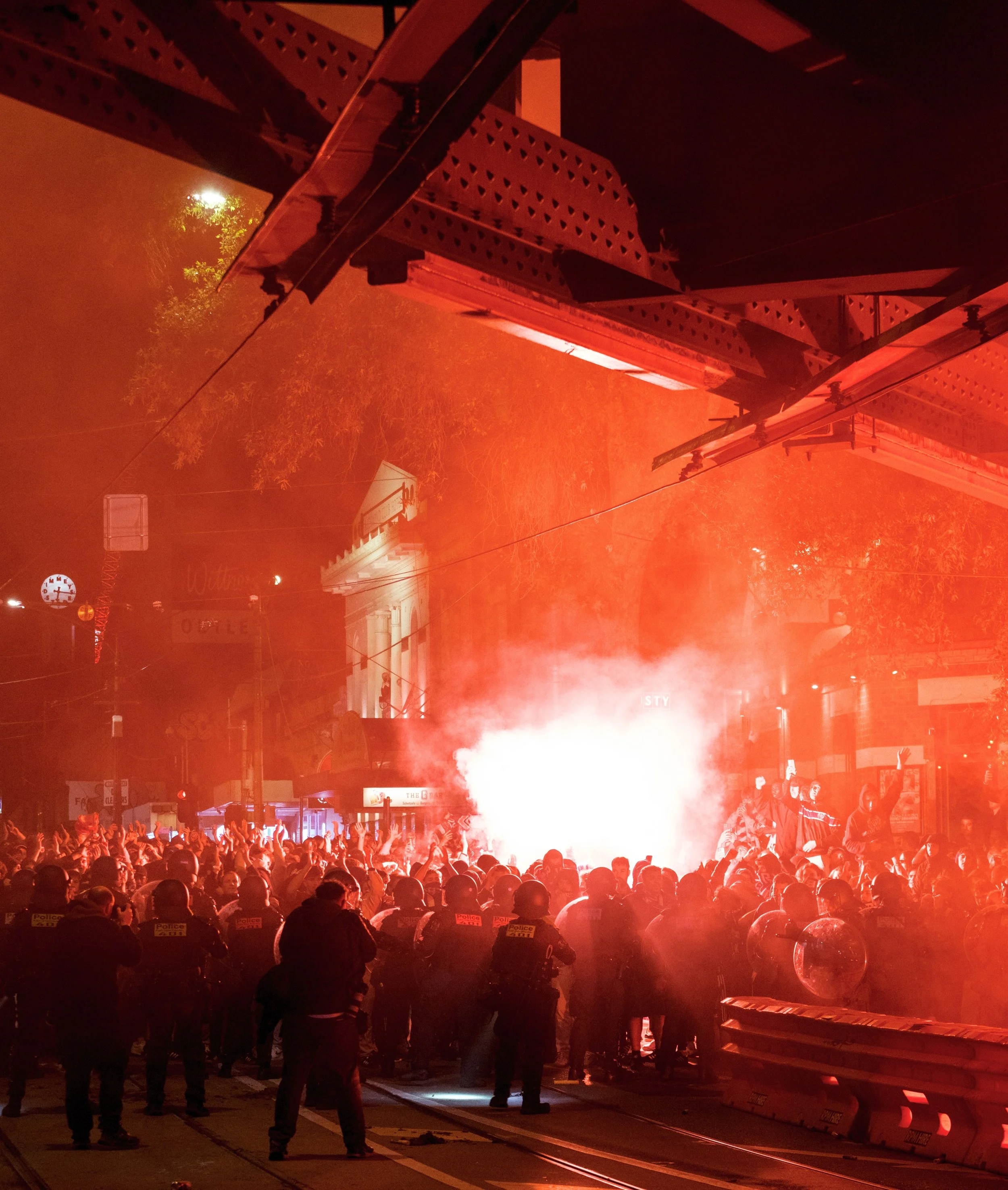 Crowd of people, including police officers, gathered on a street at night with large flames and smoke in the background, creating a red glow in the scene.