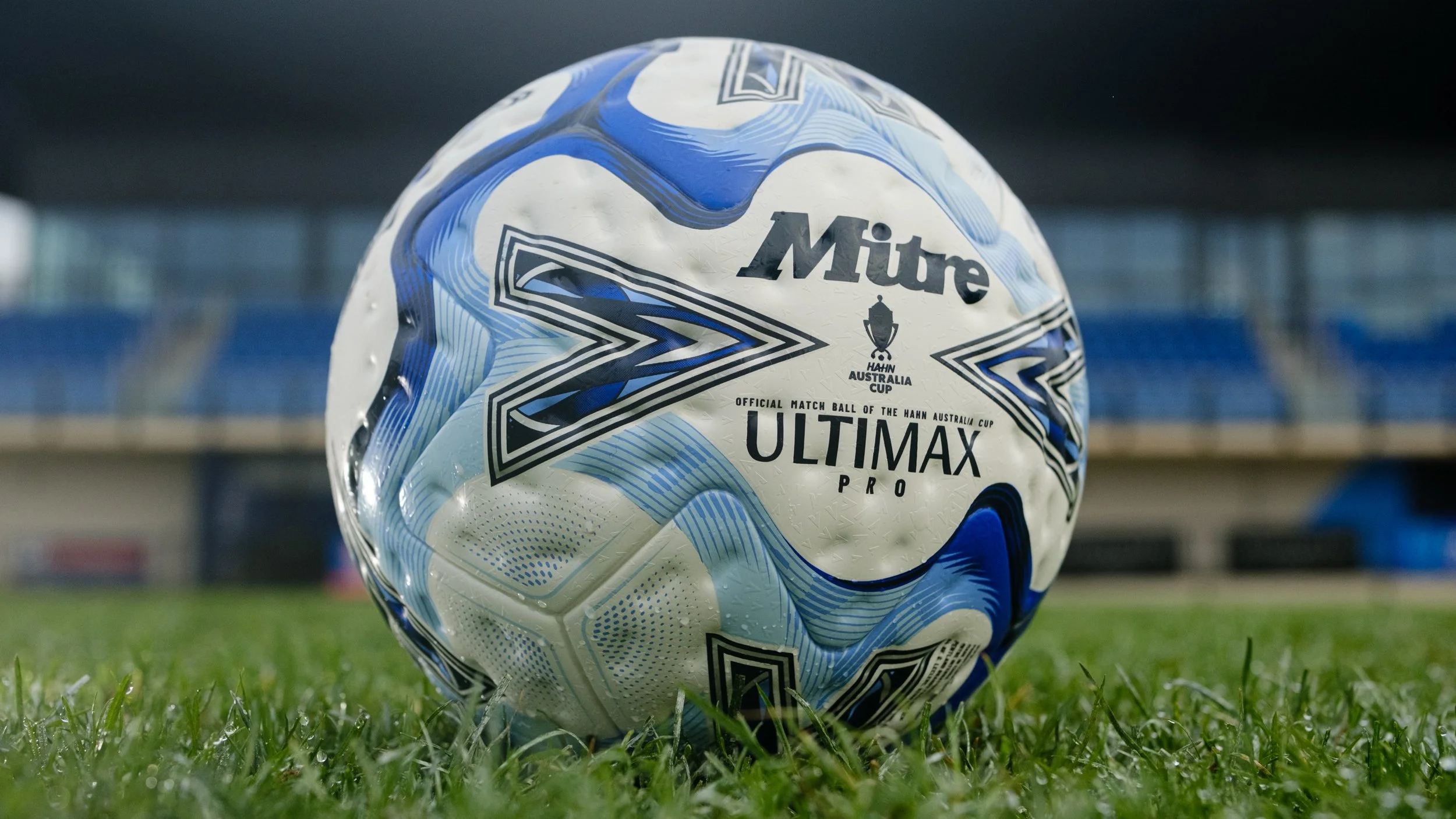 A white and blue Mitre Ultimax Pro soccer ball on grass in a stadium.