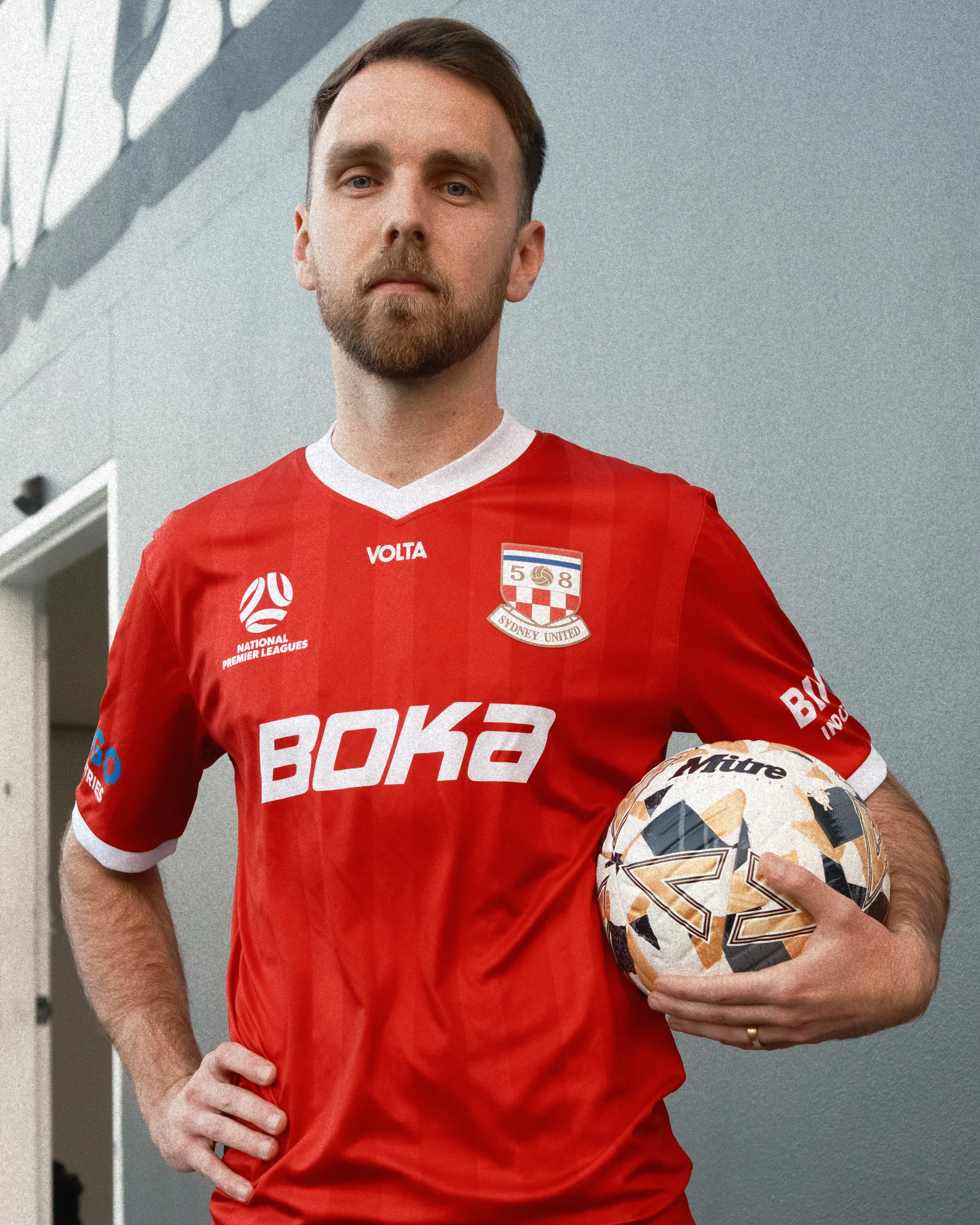 A man in a red sports jersey holding a football, standing indoors against a light gray wall.