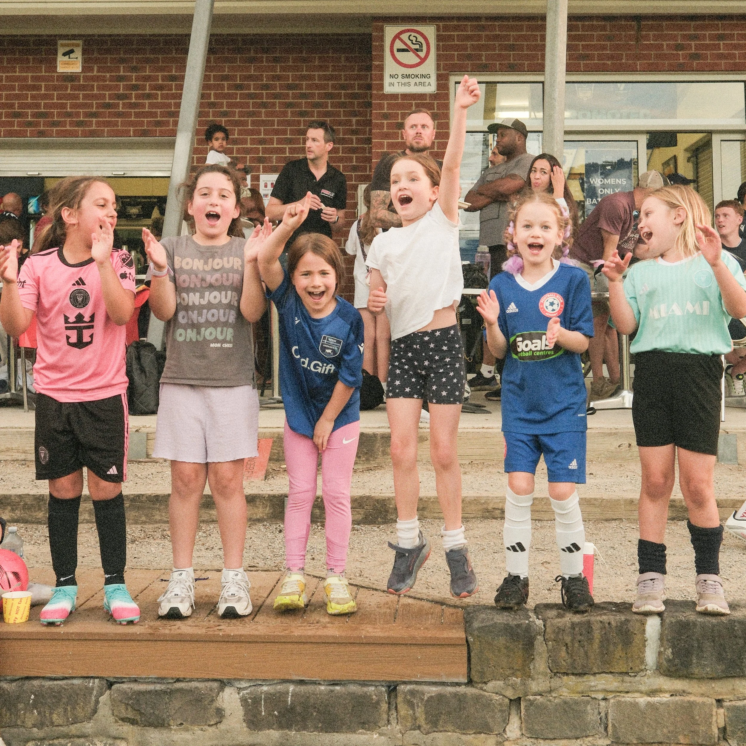 Group of young girls in sports jerseys standing on a wooden platform, some cheering, with adults watching in the background outside a brick building.