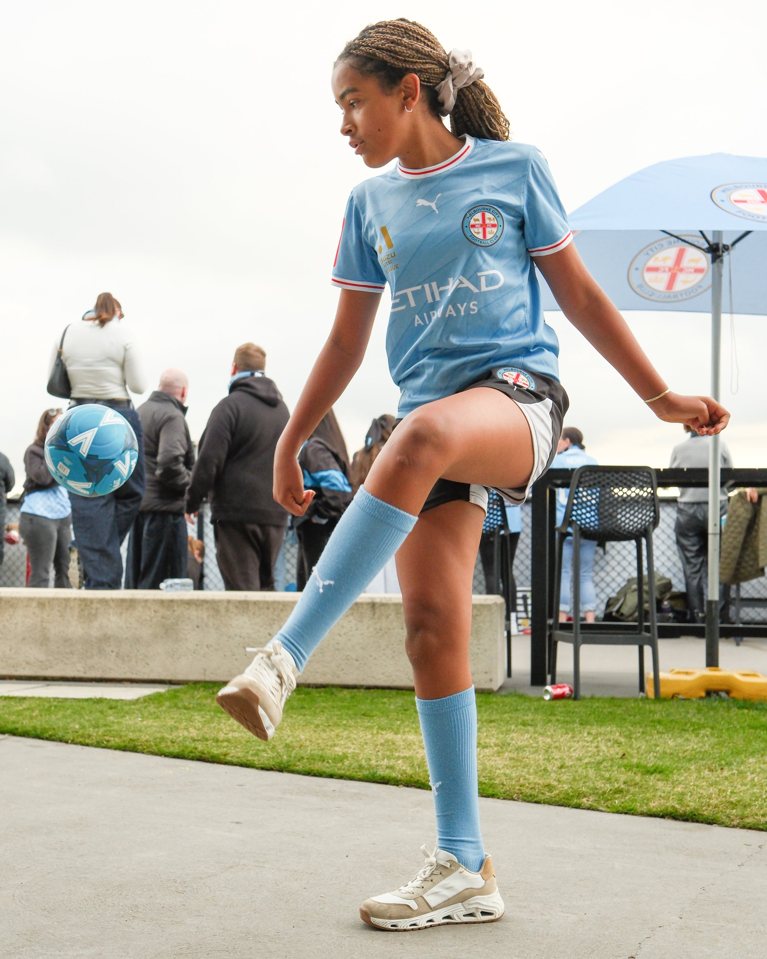 A woman in a light blue sports jersey with the logo of Melbourne City Football Club, wearing matching socks and white sneakers, is stretching her leg at an outdoor sports event. There are people and an umbrella with the club's logo in the background.