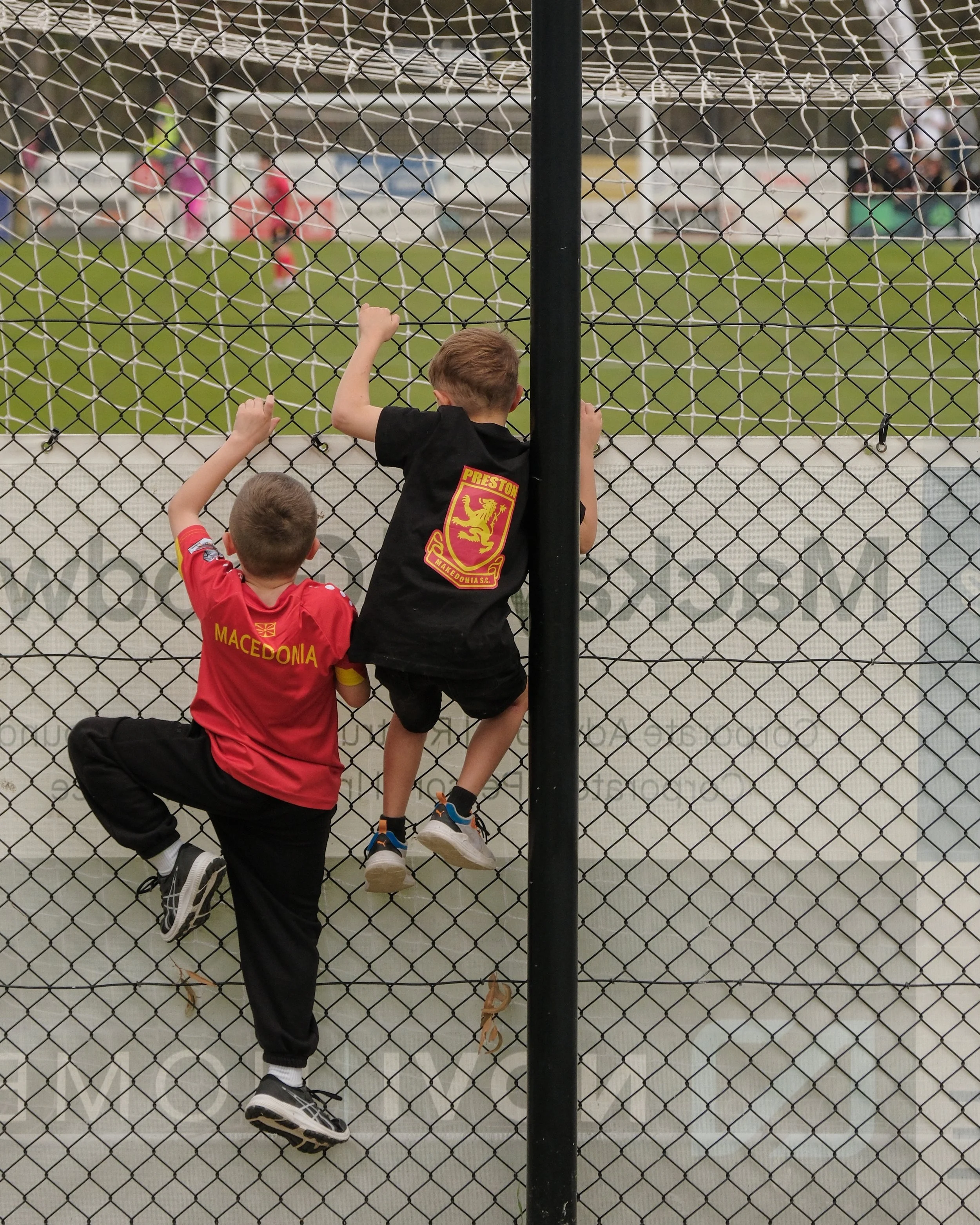 Children watching a soccer game behind a chain link fence at a sports field, with some of them sitting and others standing.