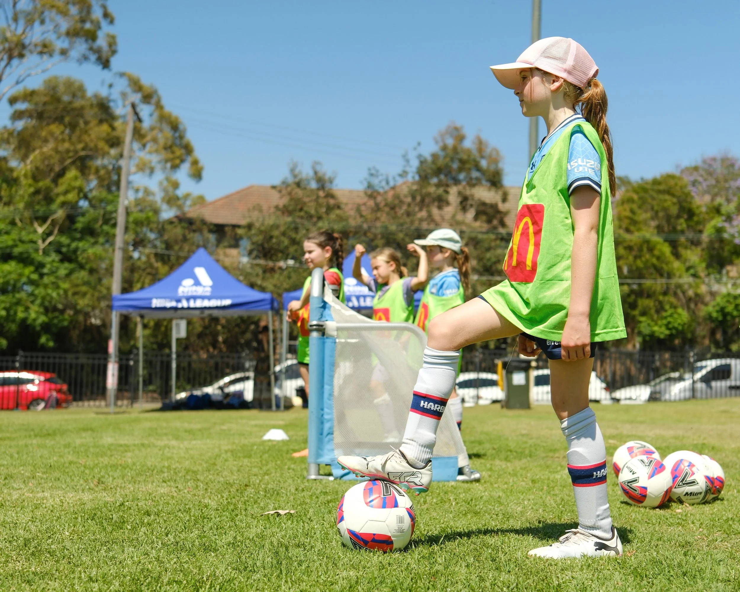 Young girl standing on a rugby field, wearing a pink cap, blue sports jersey, and green vest with a logo, lined up with soccer balls at her feet, preparing for a game or practice session on a sunny day.