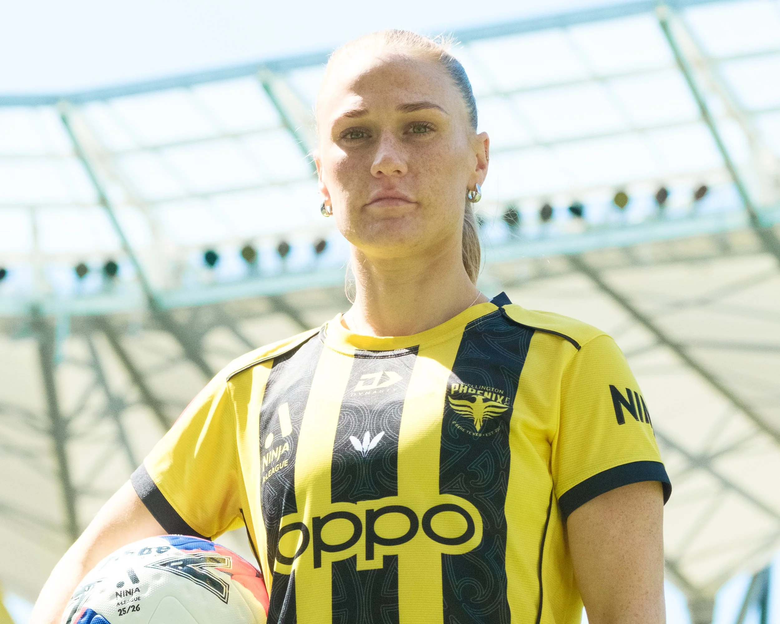 Close-up of a female soccer player with blonde hair tied back, wearing a yellow and black jersey with Nike and Adidas logos, holding a soccer ball, on a sunny outdoor sports field.