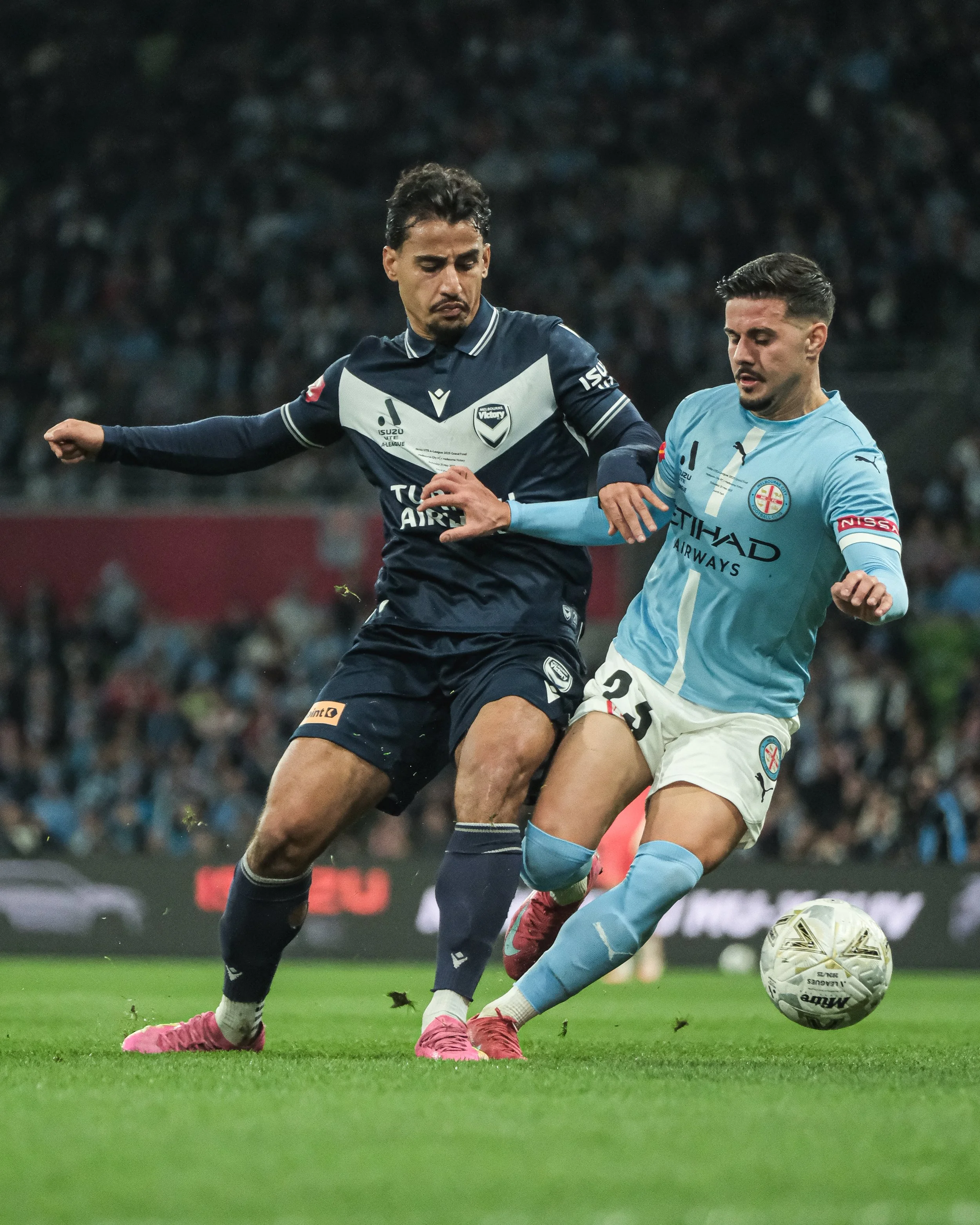 Two soccer players competing for the ball on the field. One player is in a dark blue jersey and the other in a light blue jersey. They are in motion, with grass and a blurred crowd in the background.