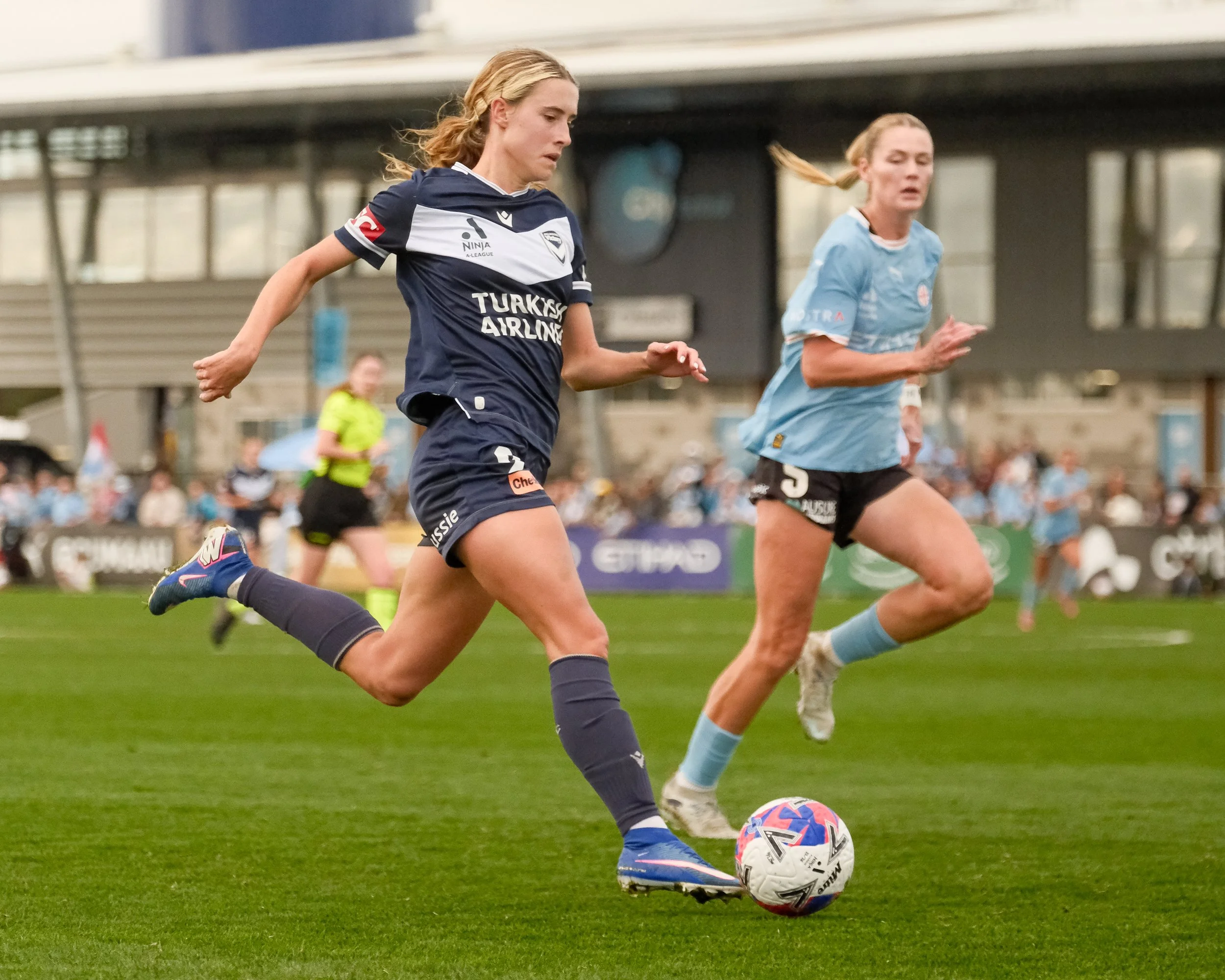 A smiling person with short blonde hair holding a colorful soccer ball above their shoulder, standing in a sports stadium with a large roof structure and empty seats in the background.