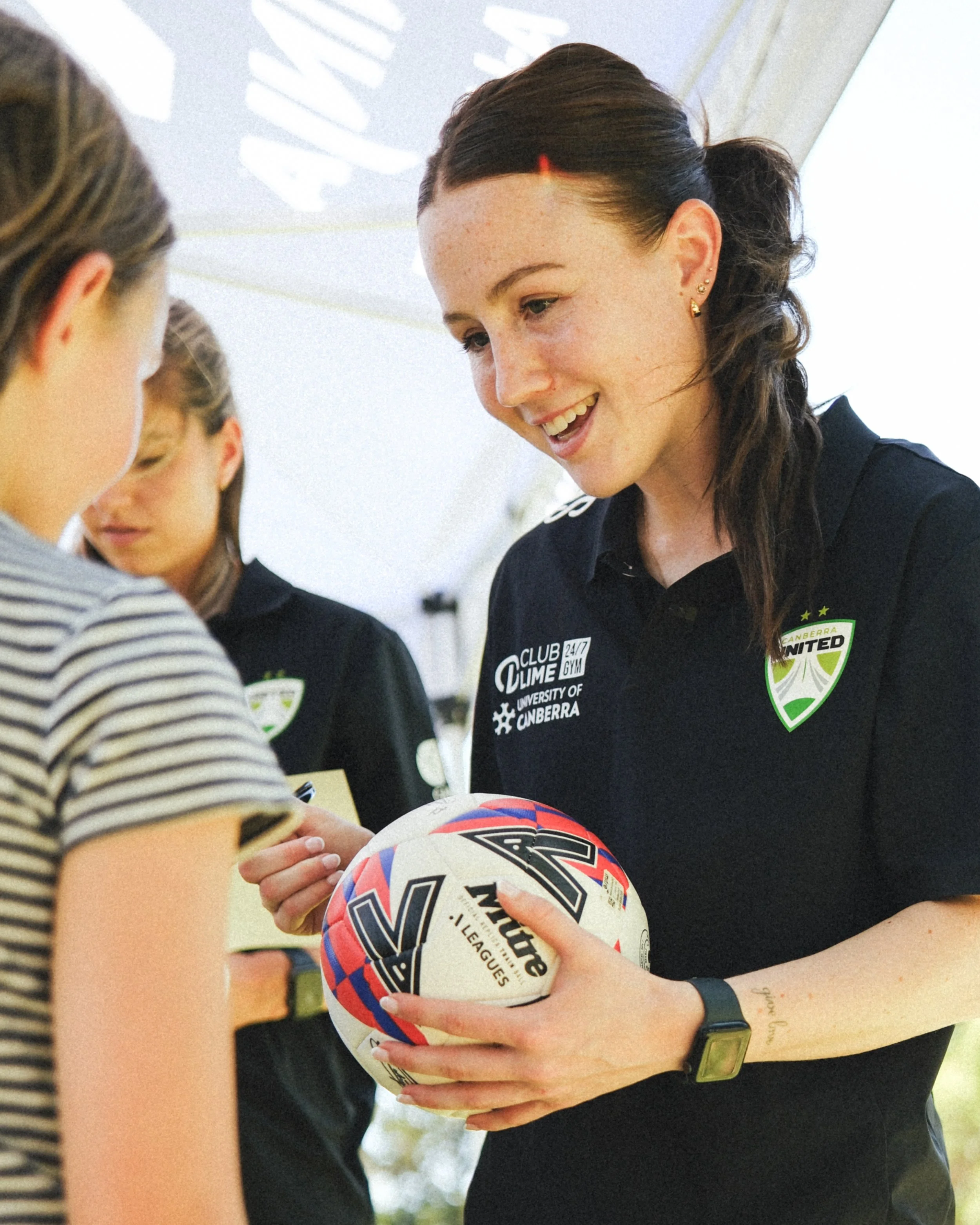 A woman with dark hair tied back, wearing a black sports shirt with various logos, holds a white rugby ball with blue and red designs, and is smiling while talking to a girl wearing a striped shirt. There are other people in the background, and the s