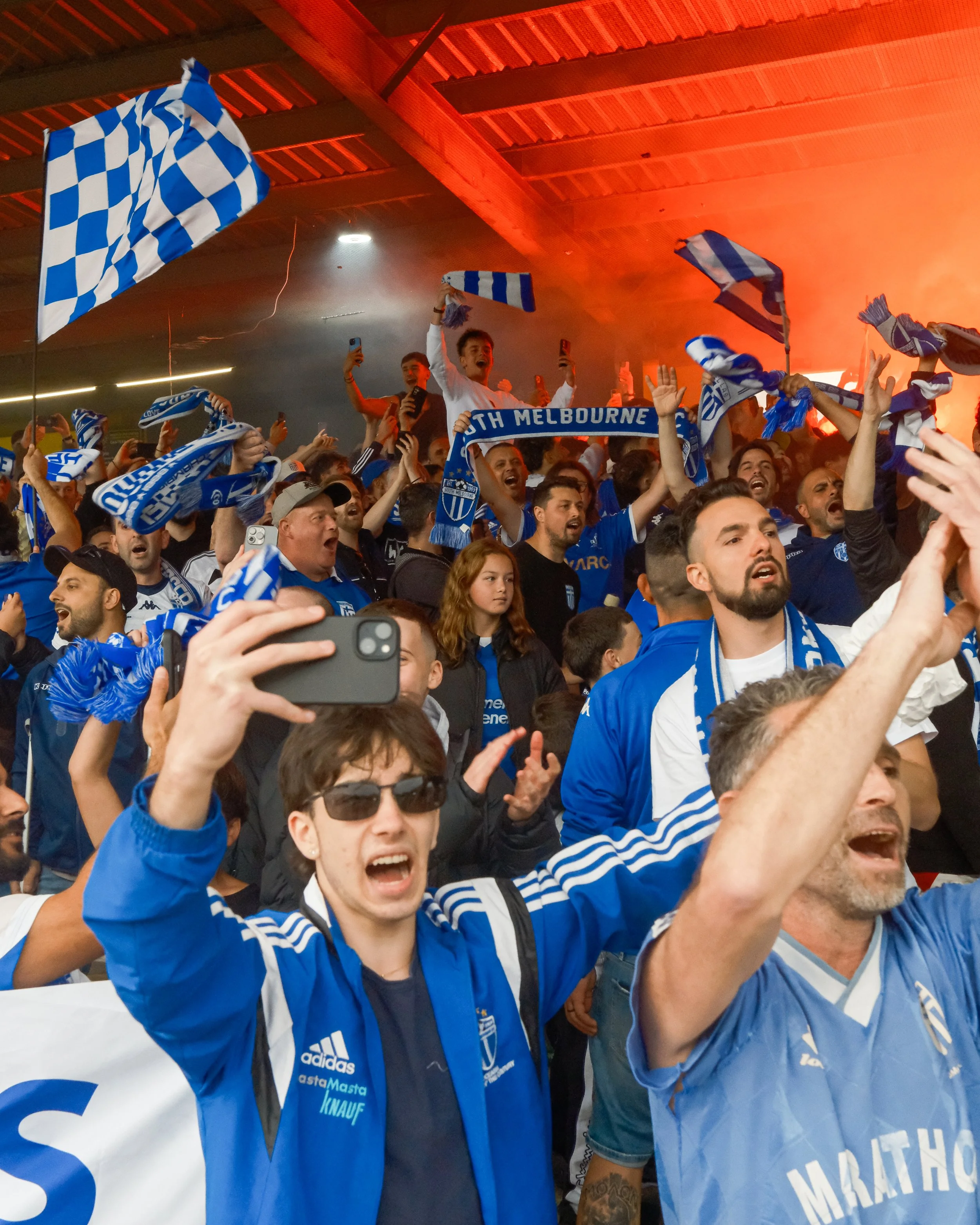 Group of enthusiastic sports fans wearing blue and white jerseys and scarves, cheering and waving flags at a stadium or indoor arena with orange lighting.