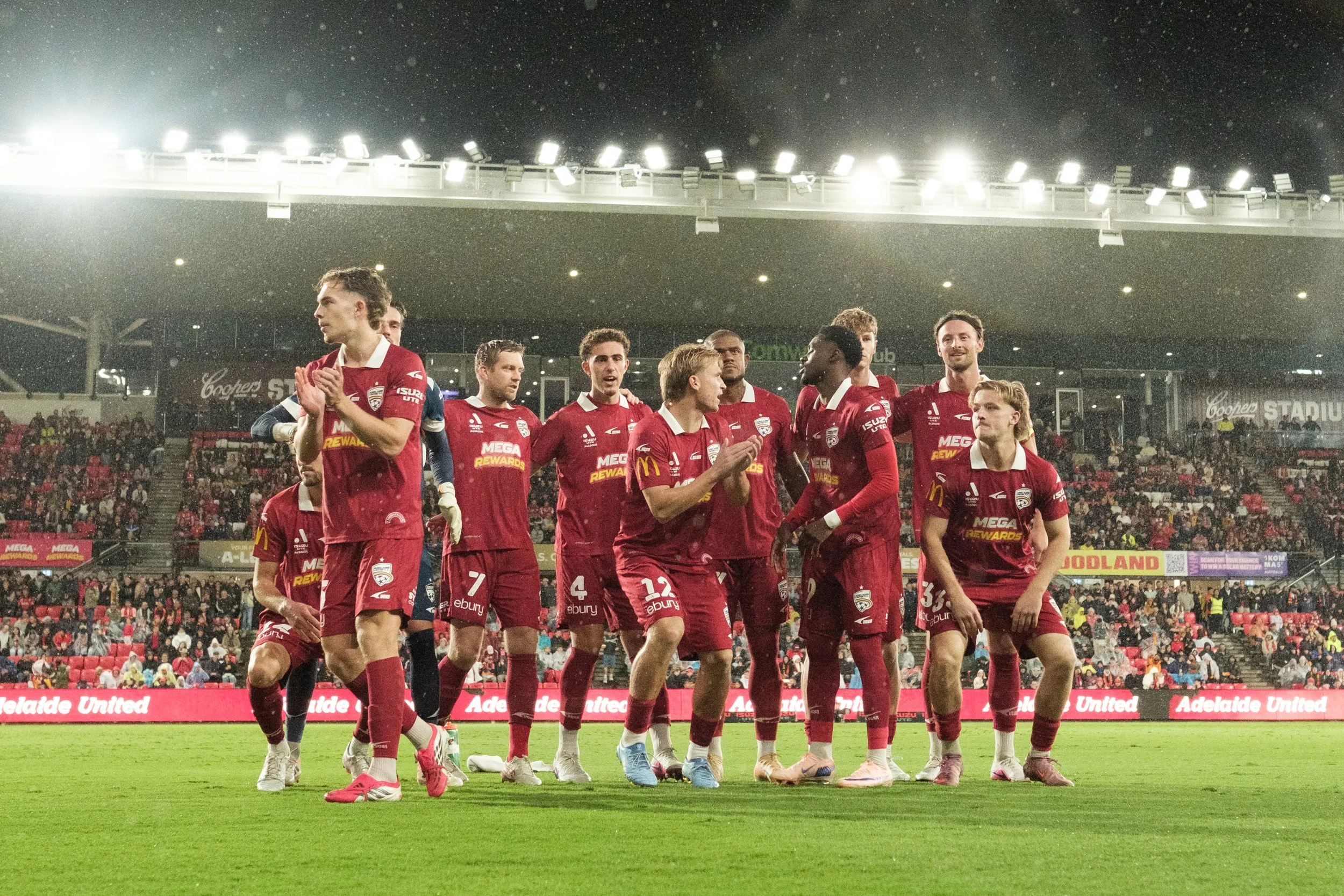 A group of soccer players in red jerseys standing on a soccer field during a match at night. They appear to be celebrating or acknowledging the crowd, with some clapping. The stadium is filled with spectators, and bright stadium lights are visible ov