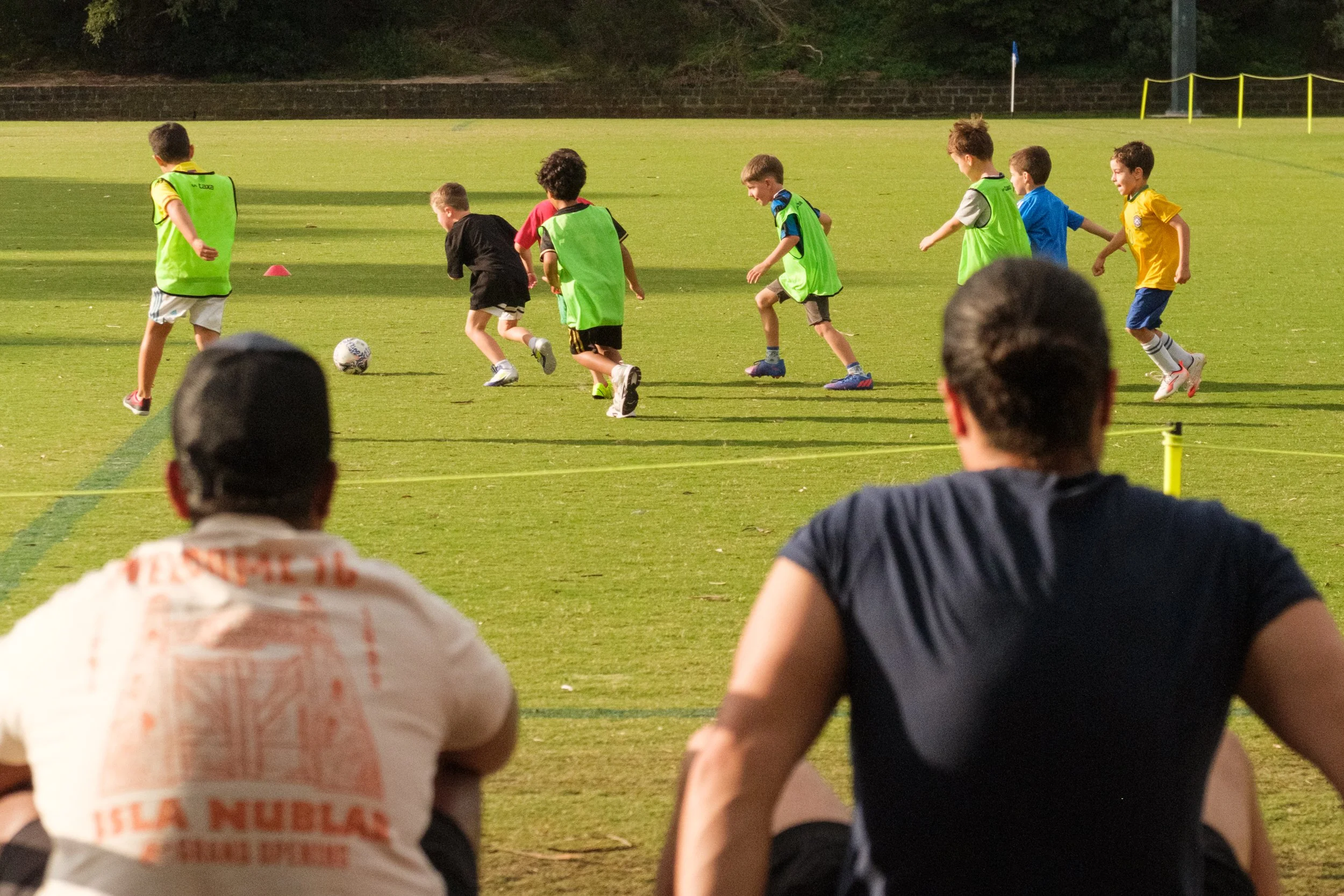 Group of children playing soccer on a grassy field with spectators watching from the sidelines during the evening.