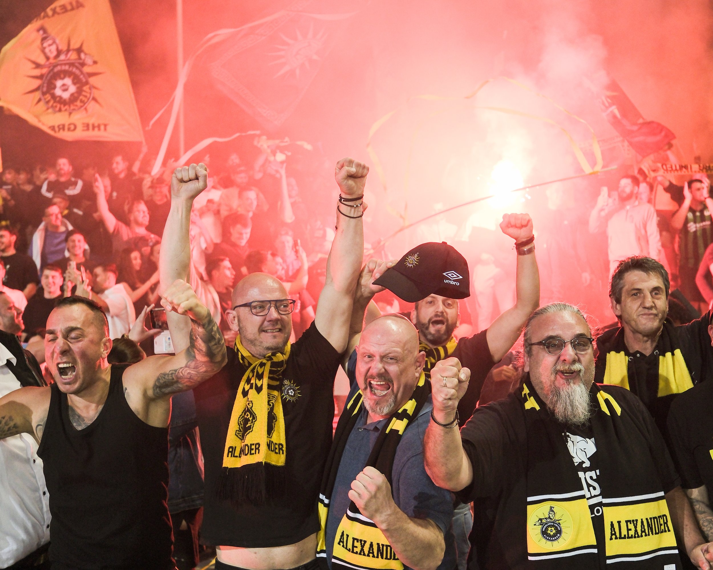 A crowd of passionate football fans celebrating, raising fists in the air, with some wearing scarves and shirts, amidst red smoke and flags in a stadium.