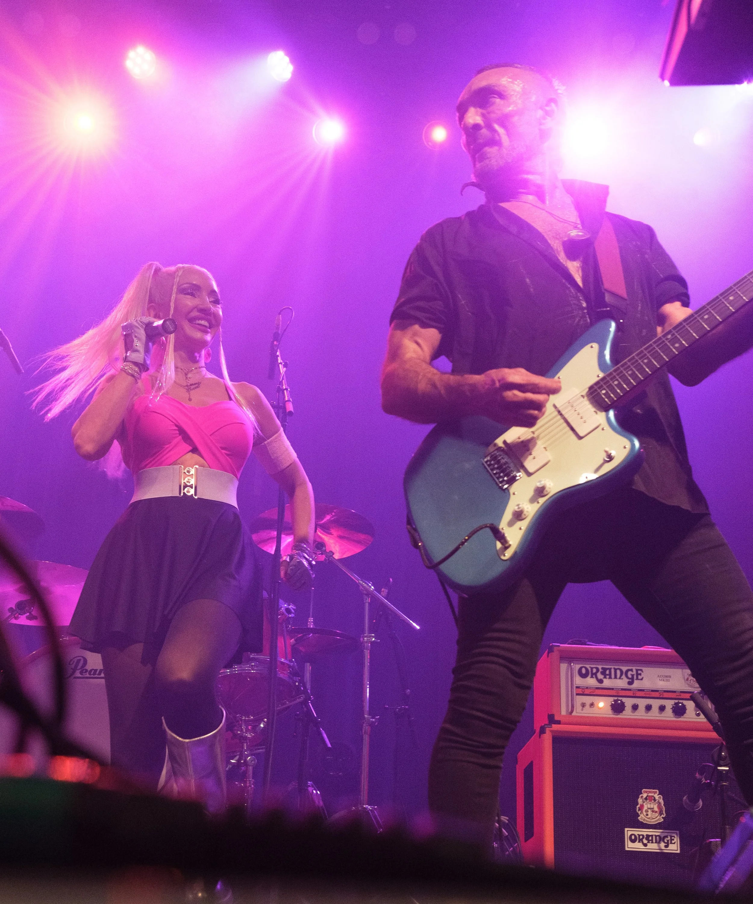 Two musicians on stage performing under purple stage lights. One woman with blonde hair, wearing a pink top and black skirt, smiling and holding a microphone. One man with short hair, dressed in black, playing an electric guitar. Musical equipment an