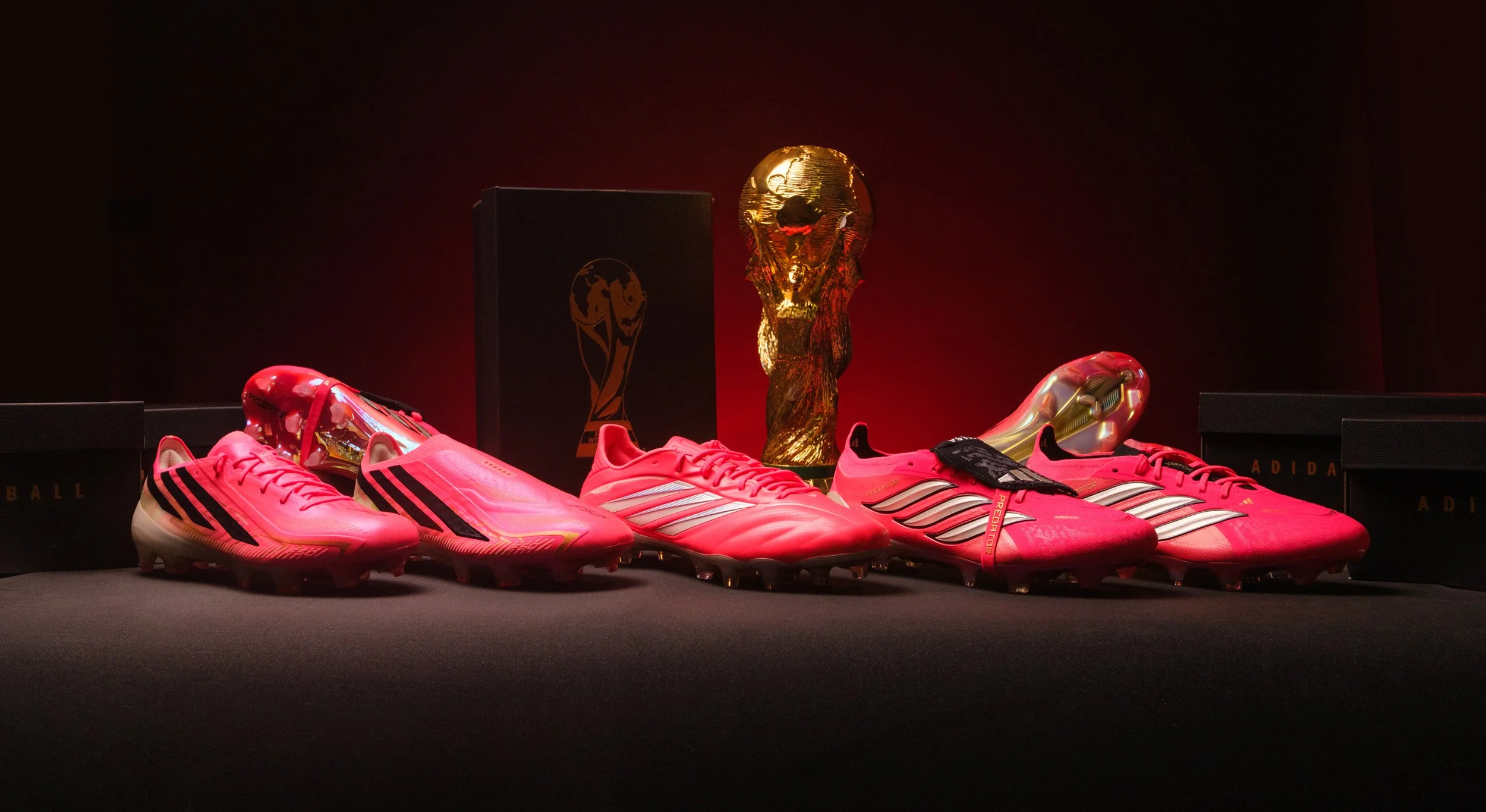 Pink Adidas soccer cleats are displayed on a black surface with a gold FIFA World Cup trophy and black box with the event logo in the background.