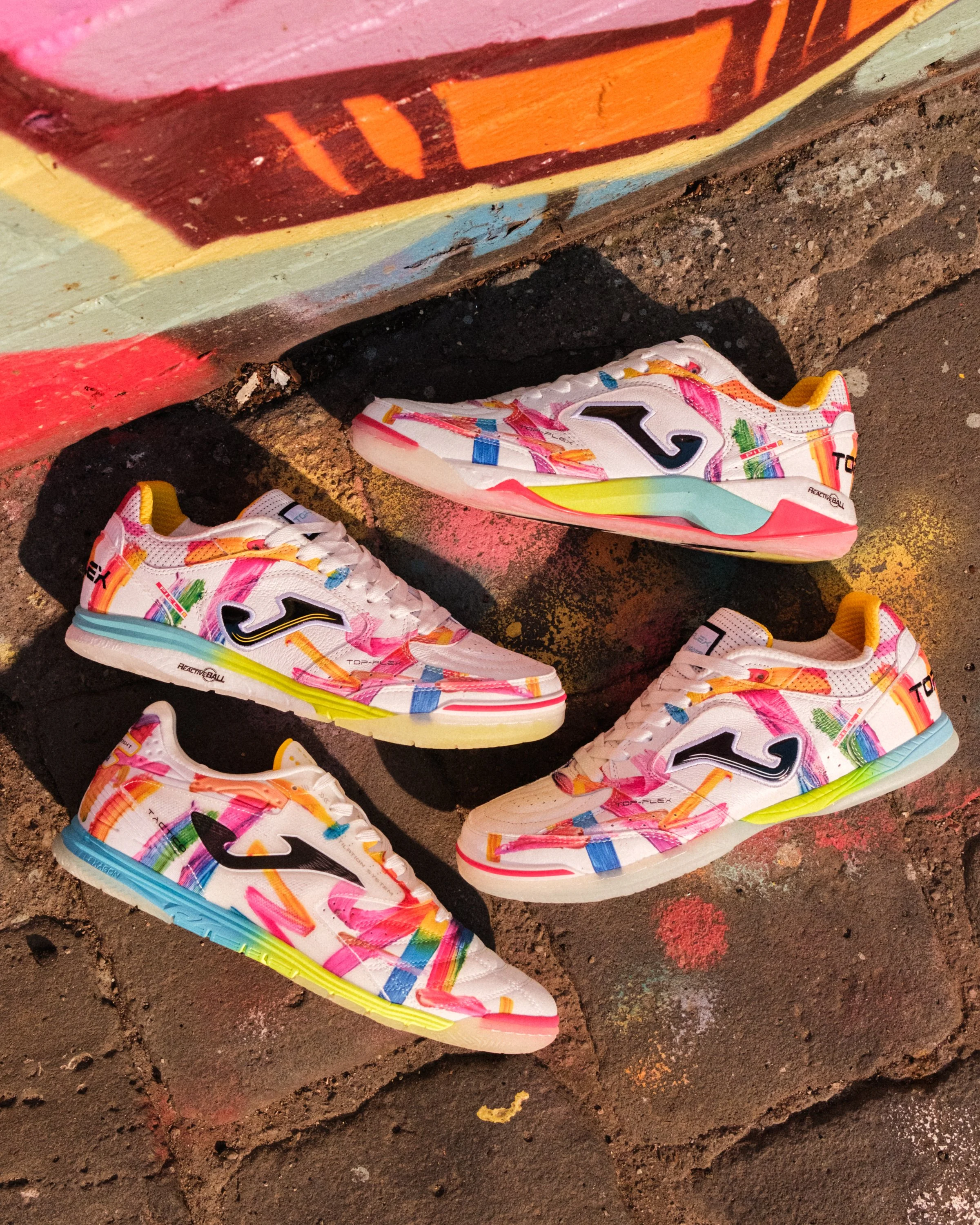 Colorful athletic shoes with a white base, decorated with bright, multicolored streaks of pink, orange, yellow, green, blue, and purple, placed on a textured, dark, and red-painted ground.