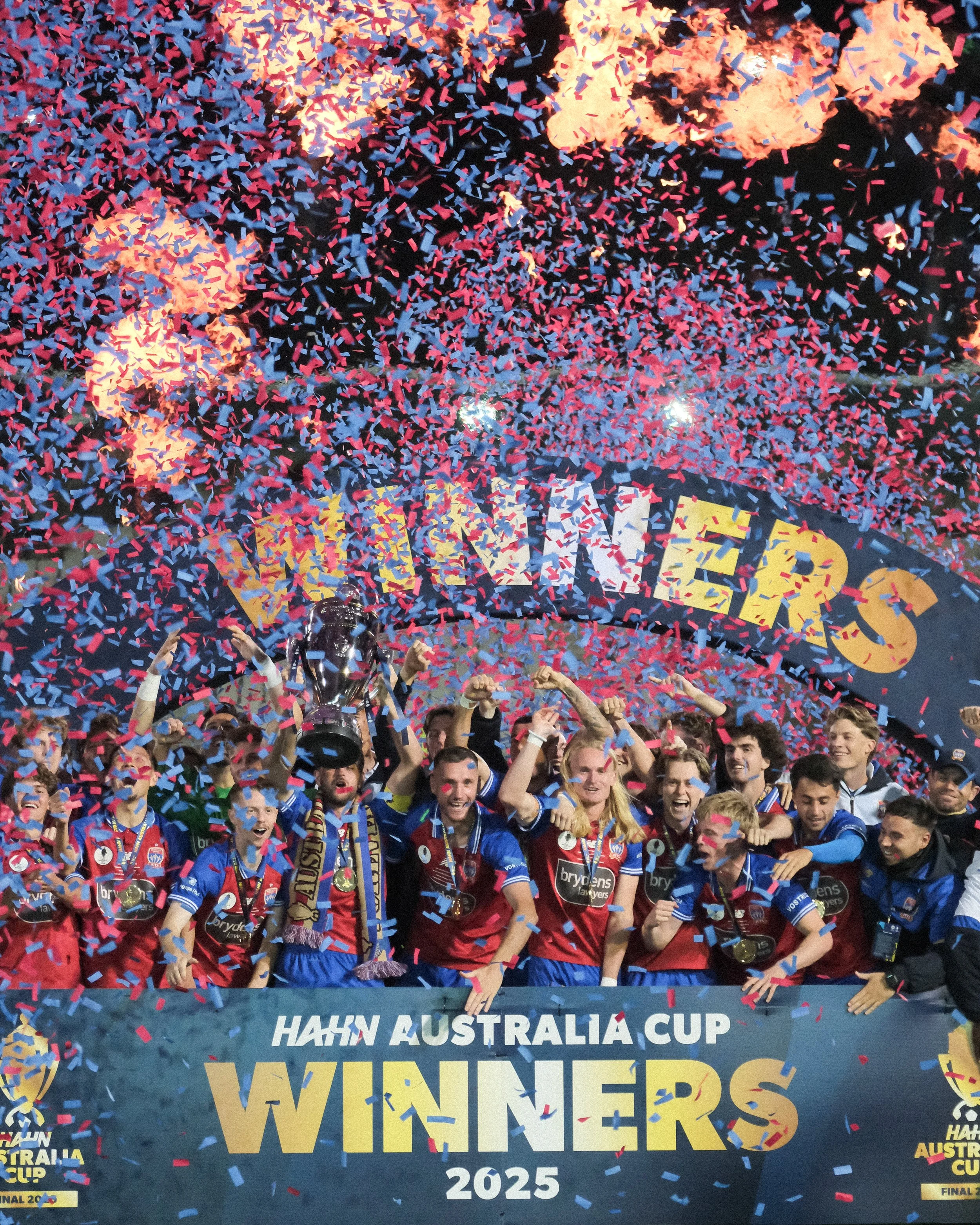 Celebrating team of players and officials holding a trophy amidst red, blue, and yellow confetti, at the Hafn Australia Cup final in 2025.