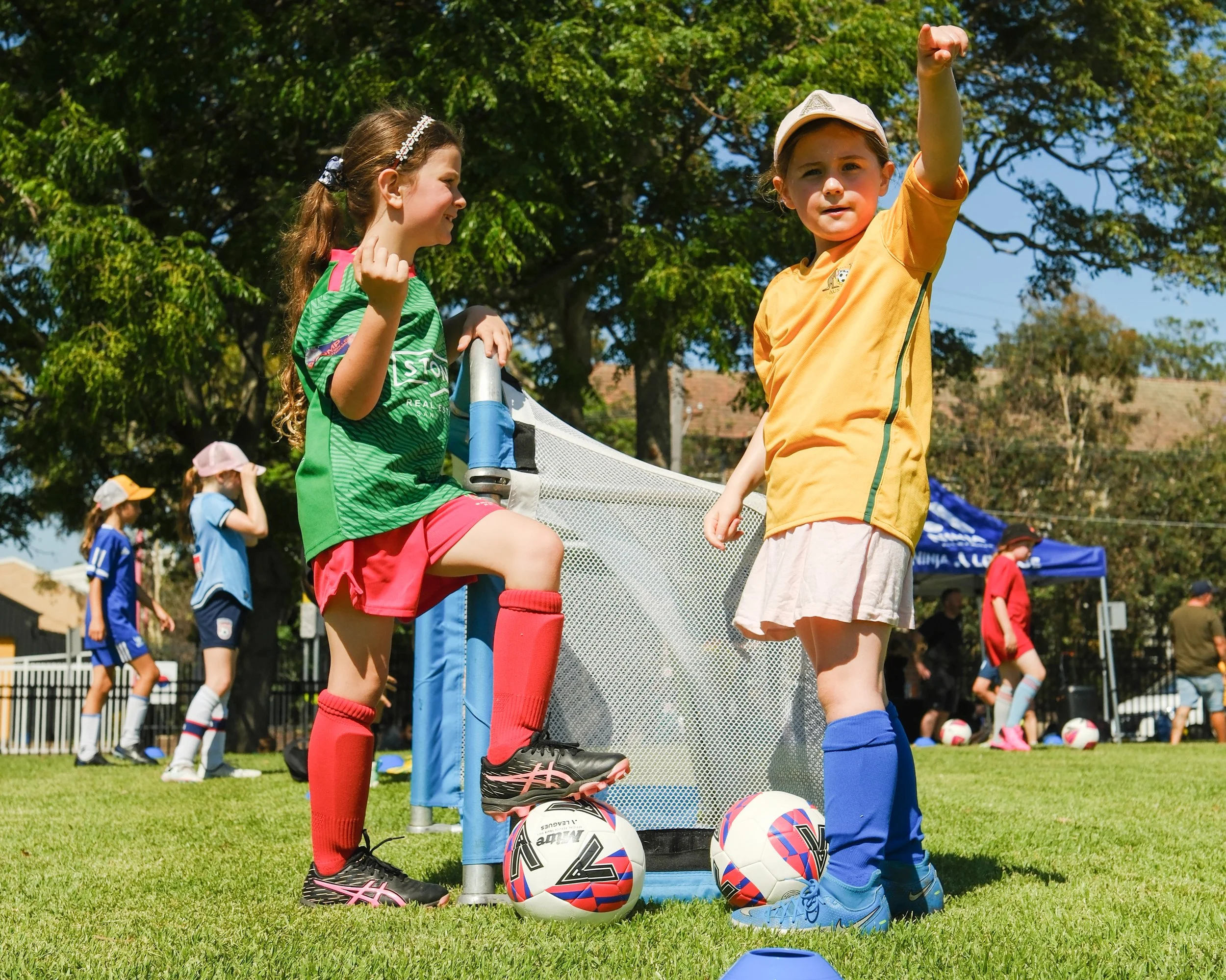 Two young girls at a soccer practice, one standing on a soccer ball with one foot, and the other preparing to kick the ball. Multiple soccer balls and cones are visible on the grass field, with other children and adults in the background, under clear
