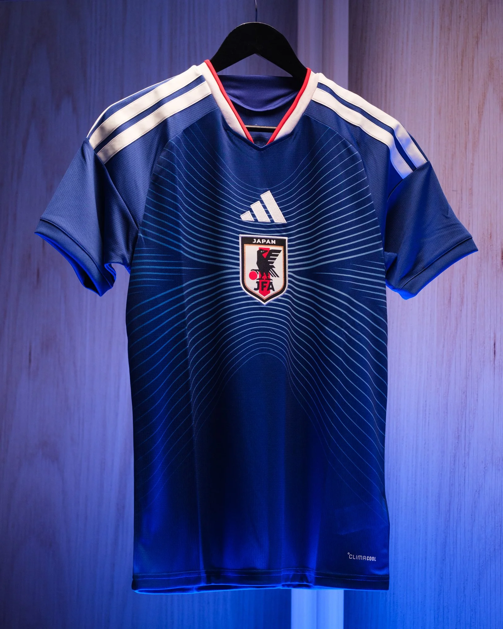 A blue sports jersey hanging on a black hanger with the Japan Football Association (JFA) logo on the front, and the Adidas logo above it. The jersey features white and light blue stripes on the shoulders and subtle horizontal and vertical lines on th