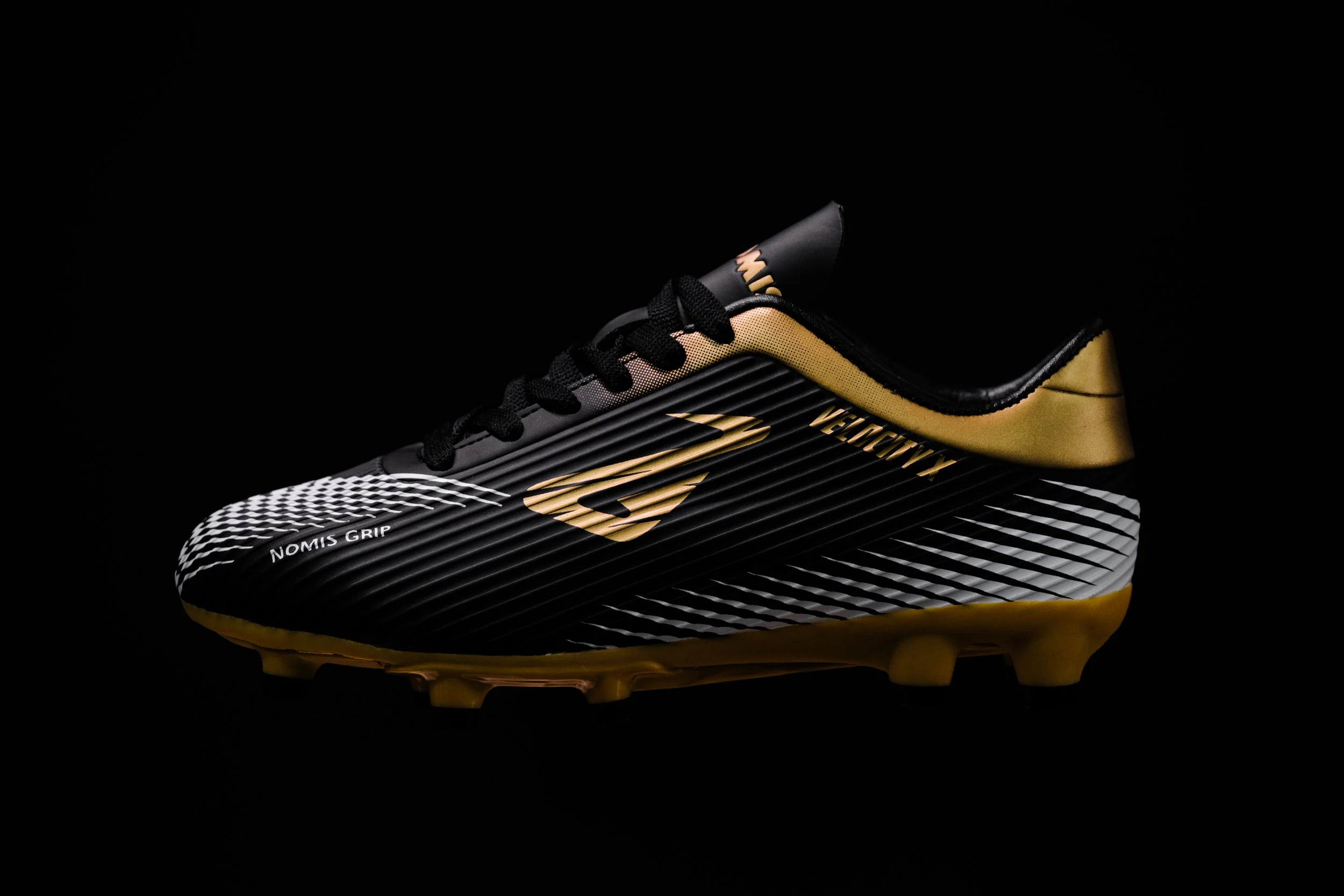 Close-up of a black and gold soccer cleat with black laces, featuring the word on the side, against a black background.