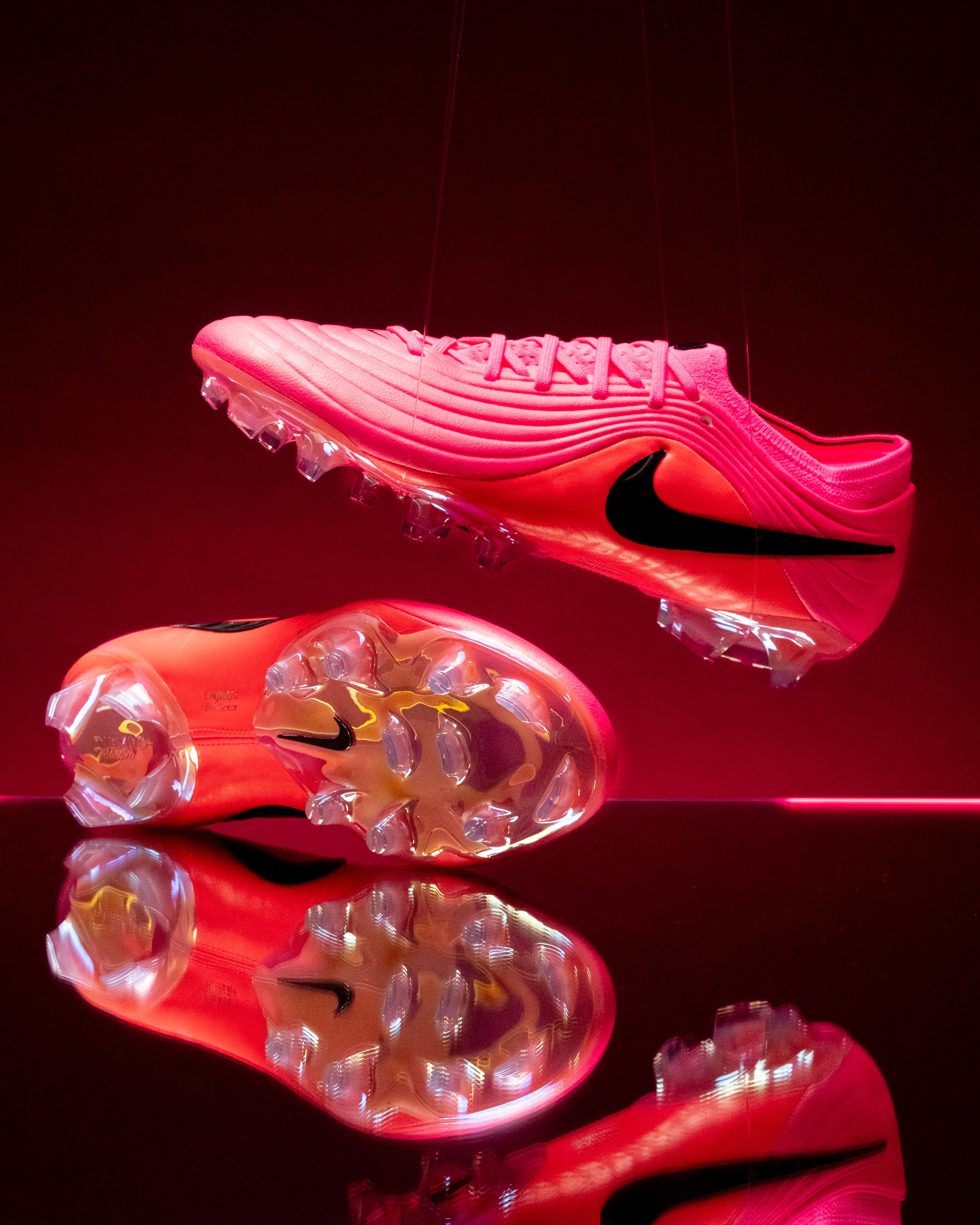 A pink Nike running shoe with a black swoosh logo displayed against a dark background. The shoe is reflected on a shiny surface below, creating a mirror image.