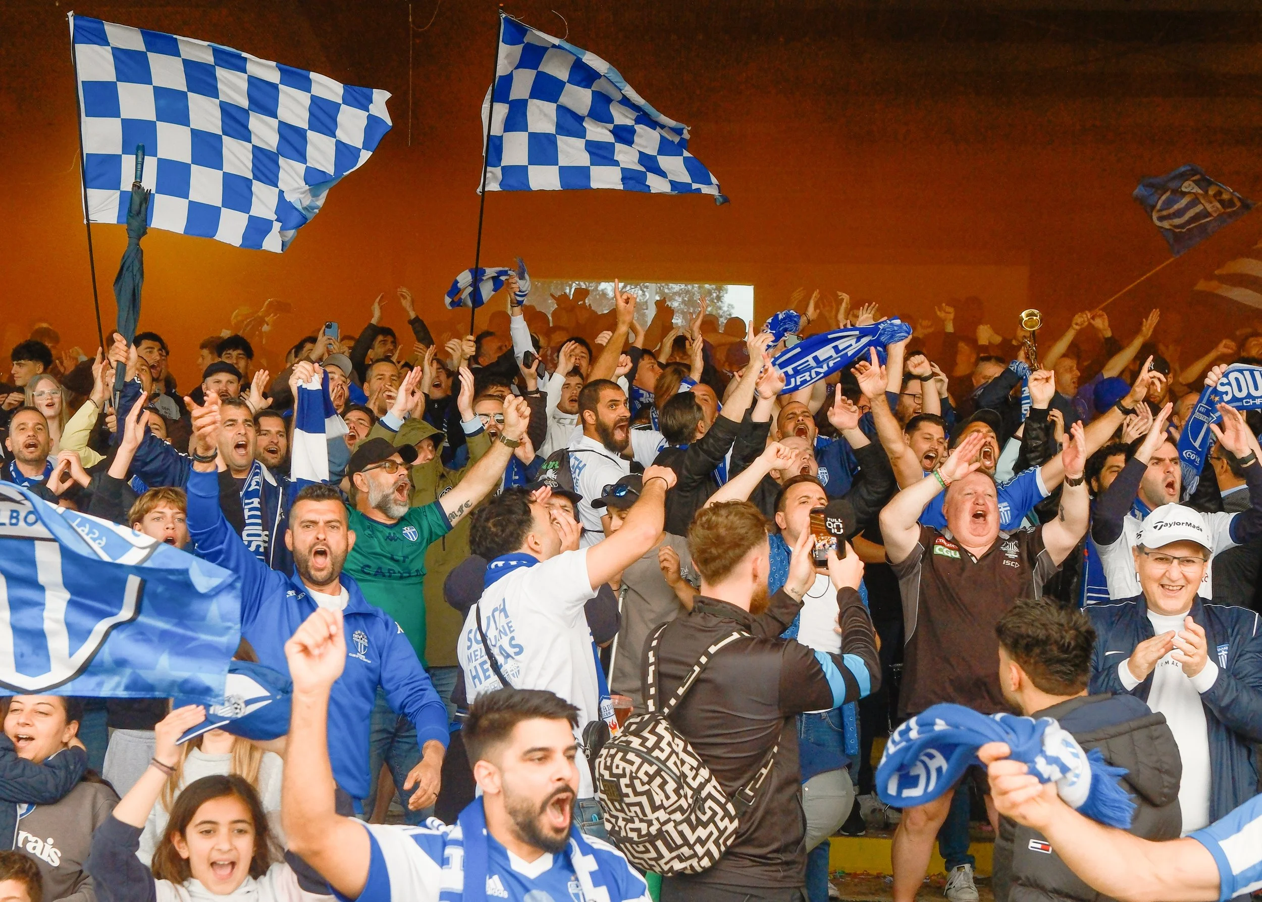 A large group of people celebrating, wearing blue and white clothing, with several blue checkered flags. The scene appears to be in an indoor stadium or venue, with many individuals raising their hands and cheering.
