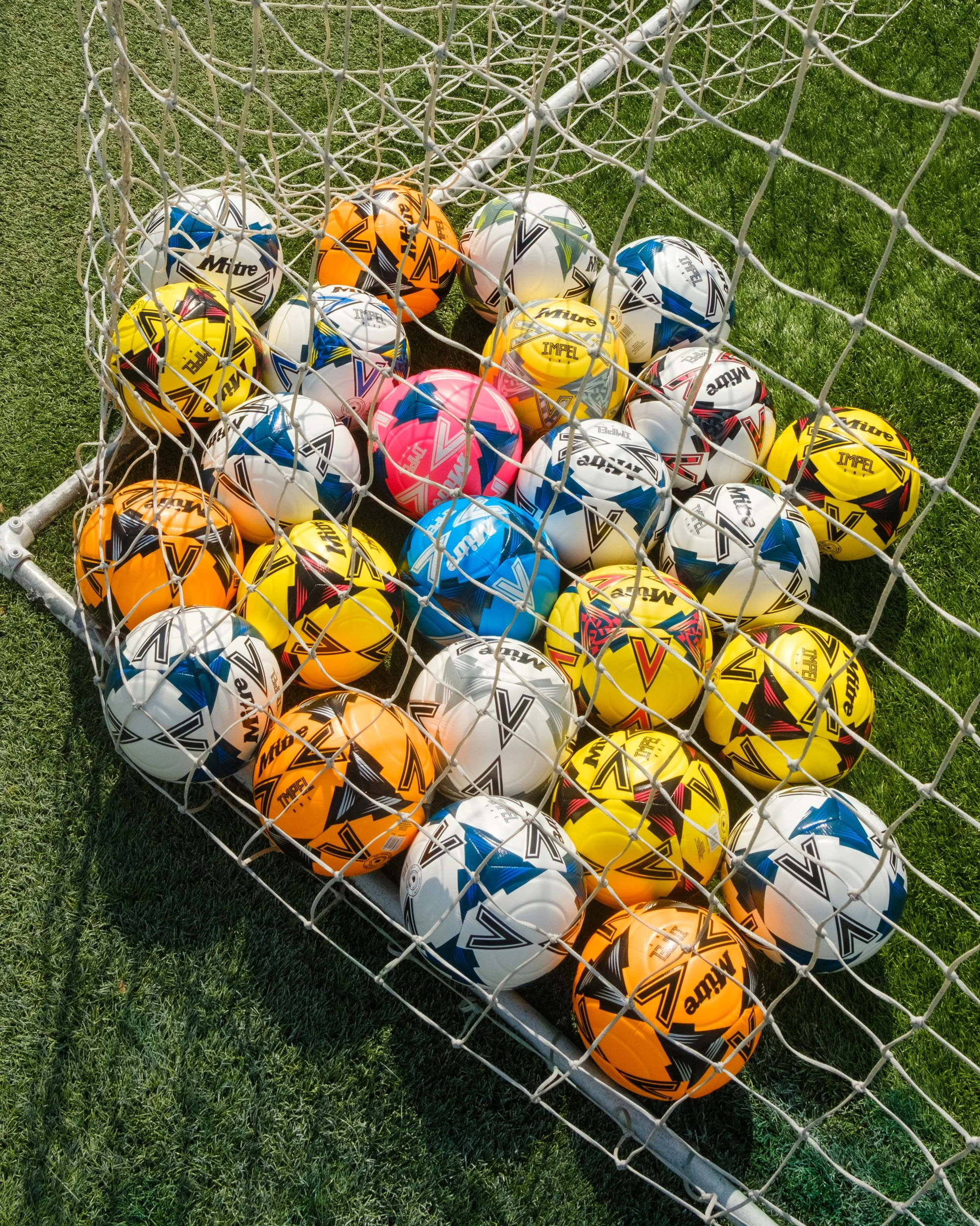 A collection of various colored soccer balls inside a soccer goal net on a grassy field.