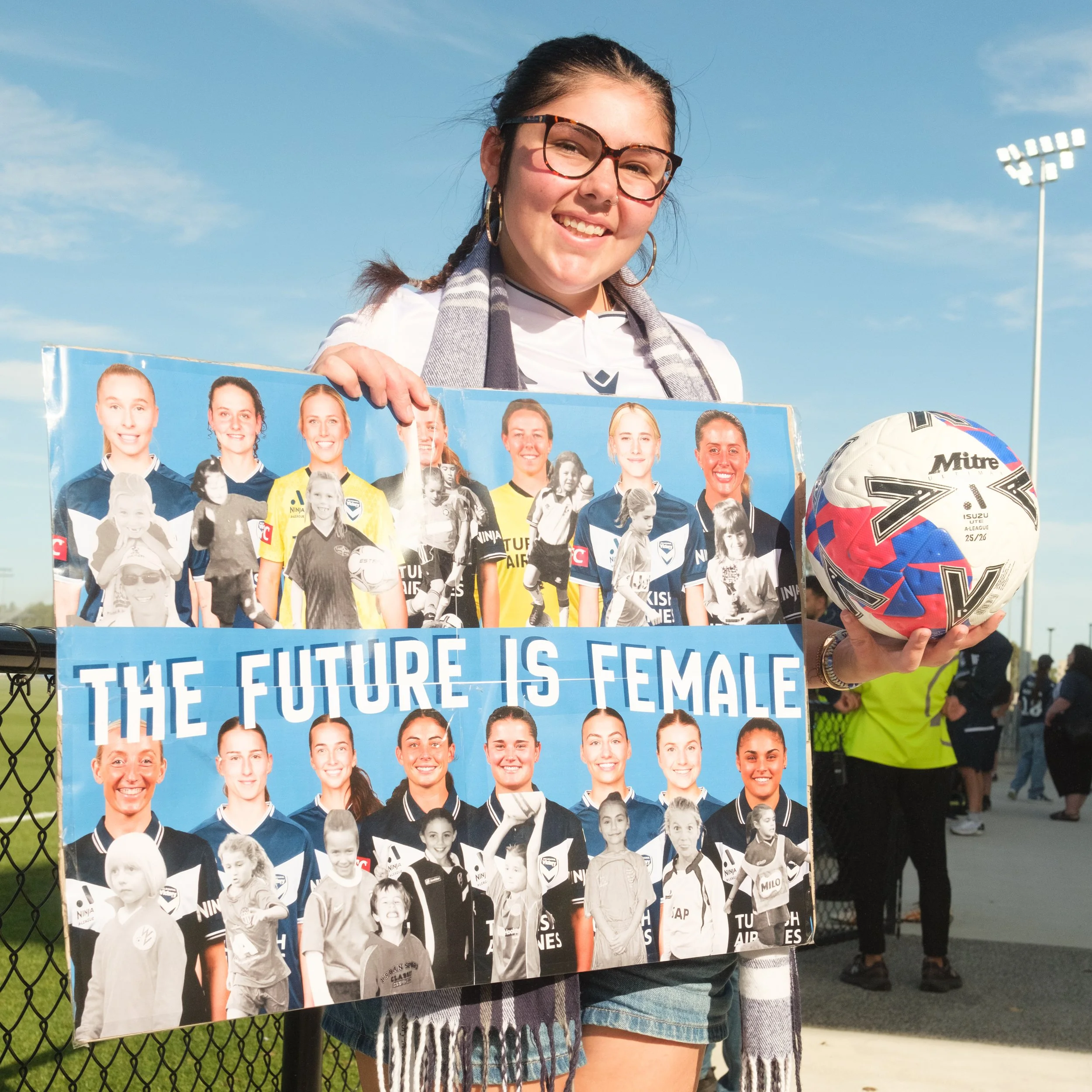 Girl holding a poster with photos of women athletes at a sports stadium on a sunny day.