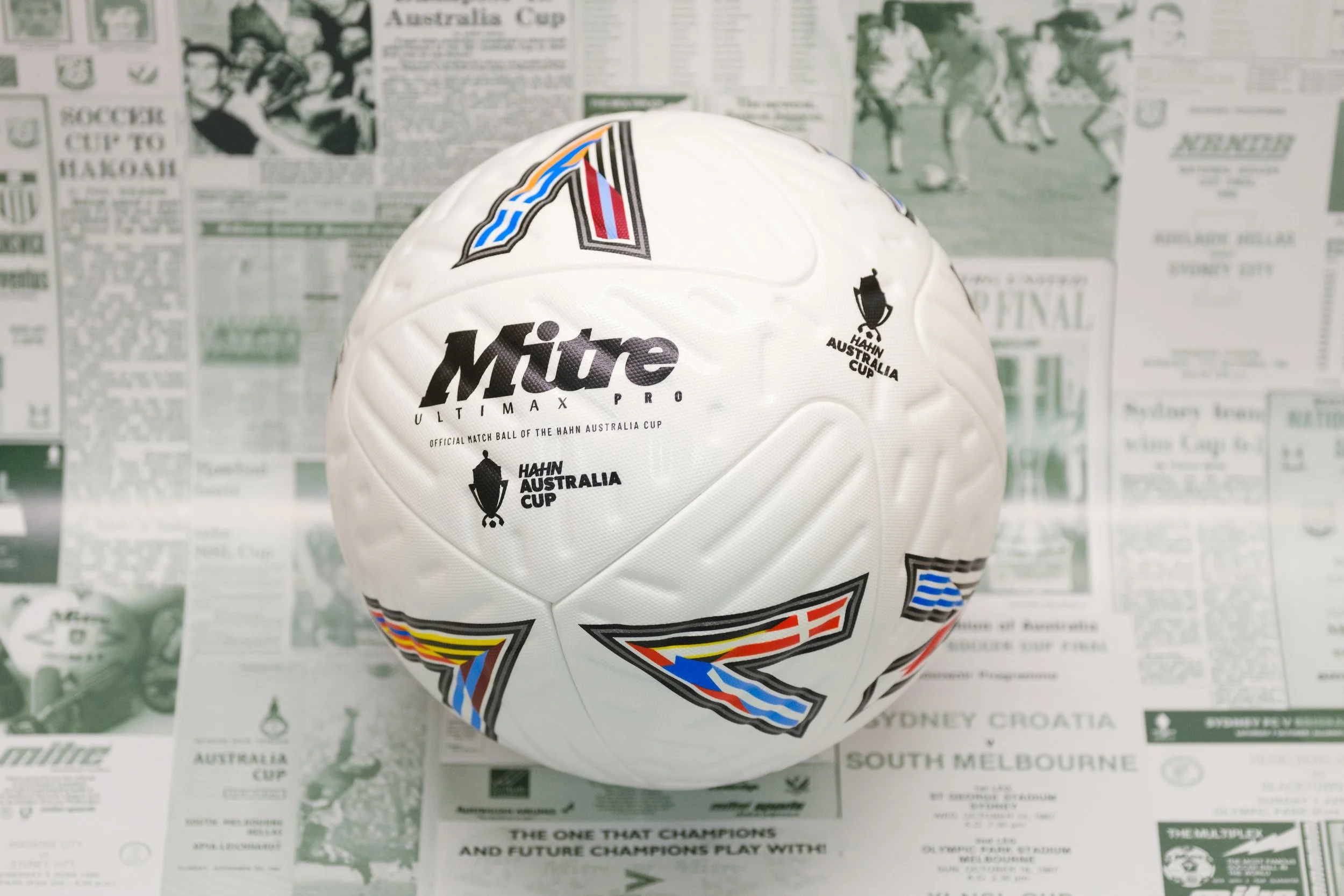A white rugby ball with logos and colorful stripes, placed on a surface covered with newspaper clippings related to sports and Australia.
