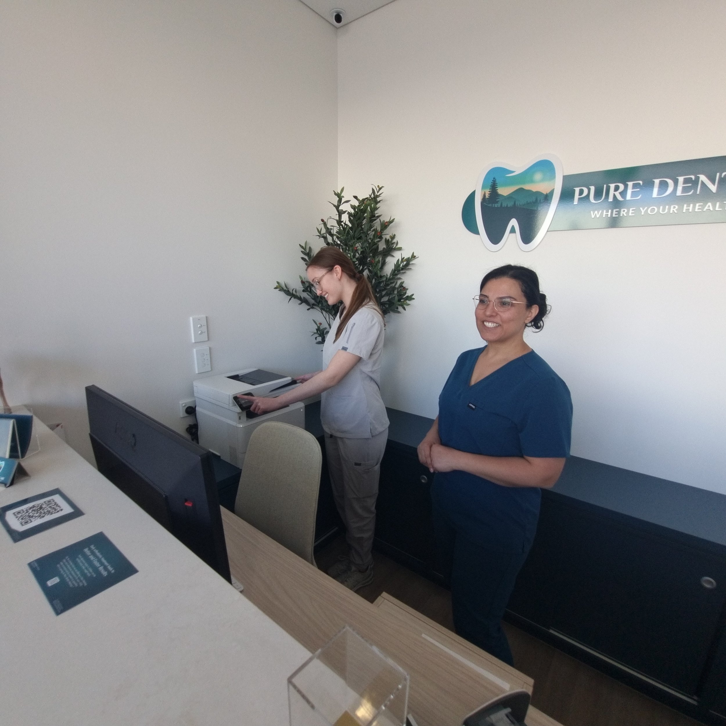 Two women standing near a wall with a sign that reads "Pure Den... where your health." One woman, wearing glasses and a dark blue shirt, is smiling, while the other woman, with long hair, is looking down at a table with a printer and informational pamphlets.