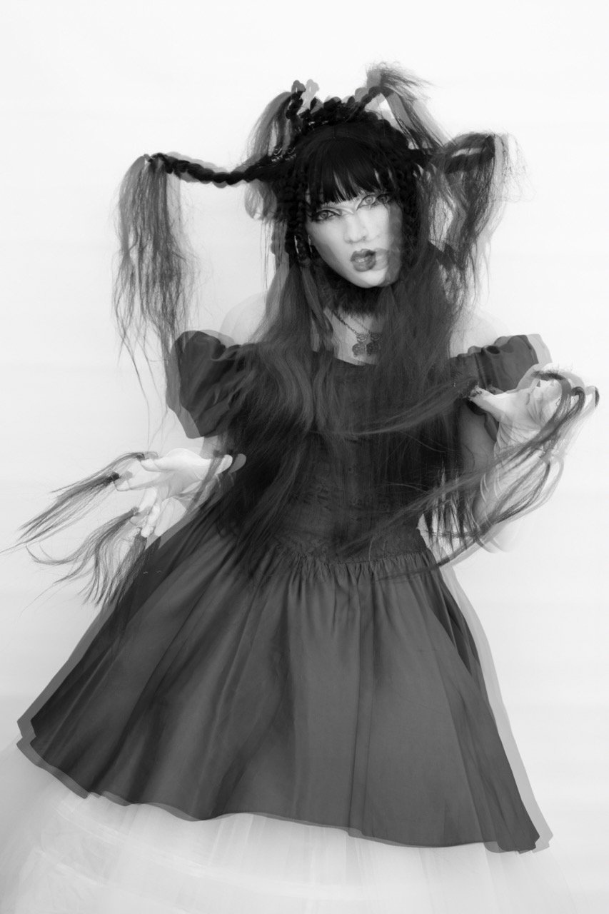 A black and white image of a person with long hair styled in braids and loose sections, wearing an off-shoulder dress, creating a ghostly effect with double exposure.