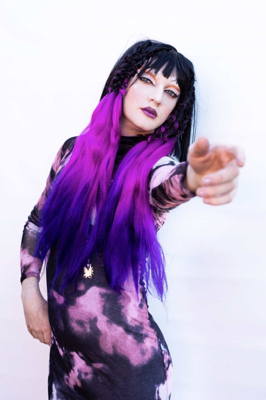 Person in a purple and black wig and tie-dye outfit, gesturing towards the camera.