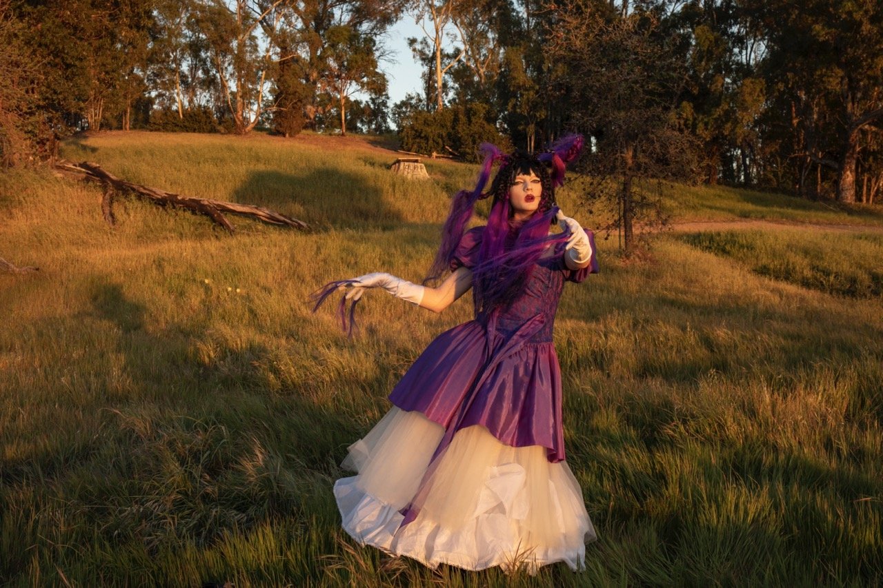 Person in elaborate purple costume with white gloves, standing in a grassy field, surrounded by trees.