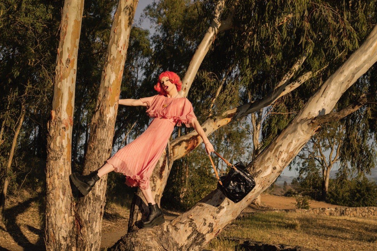 Person with pink dress and red wig poses on tree with black boots and handbag in a forest setting.