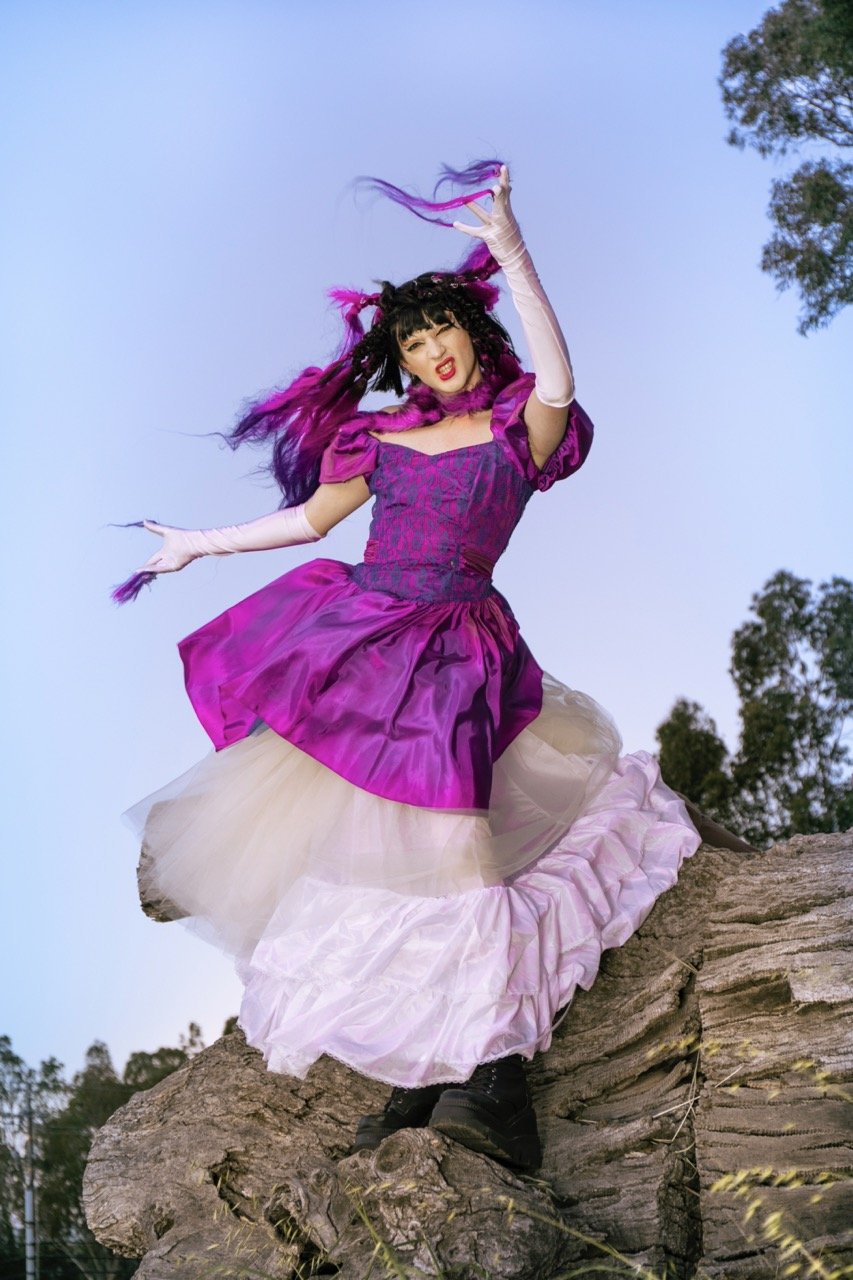 Person in a vibrant purple and white costume with feathered headdress, posing on a rock, against a clear blue sky.