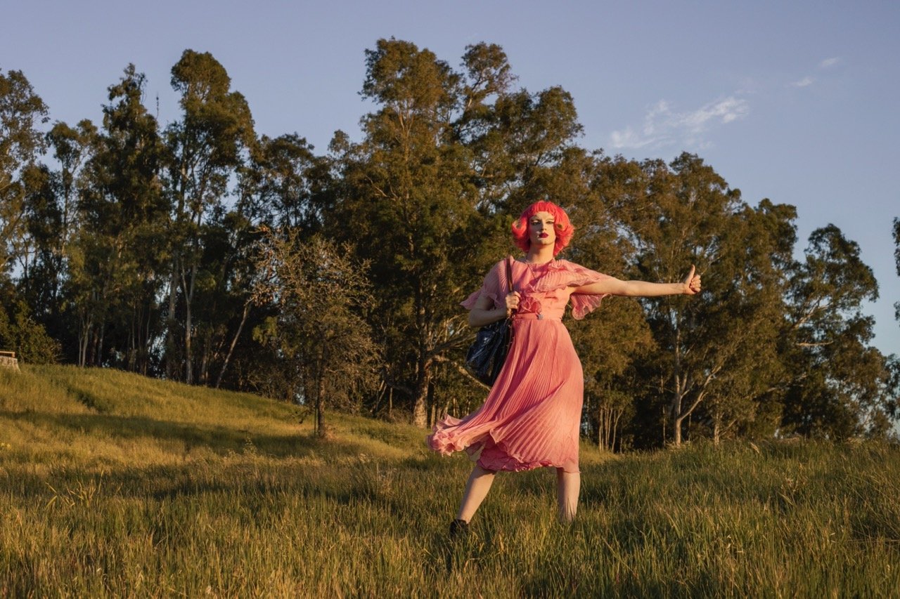 Person with pink hair in a dress standing in a grassy field with trees in the background.