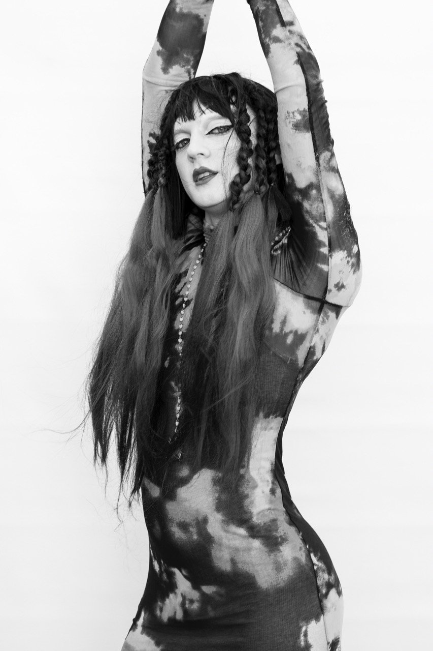 Black and white photo of a person with long hair, wearing a patterned outfit, and raising their arms above their head.