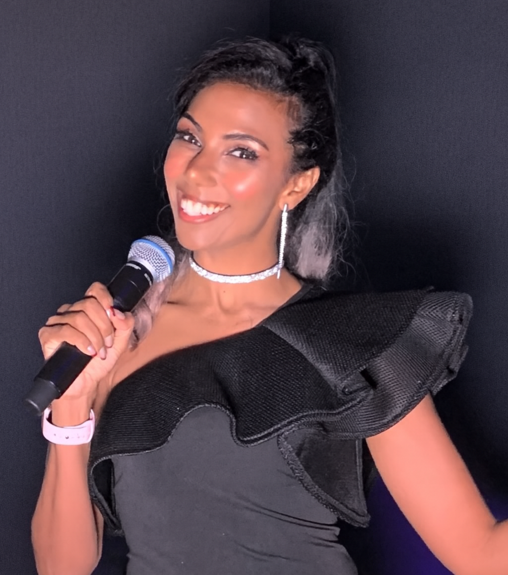 A woman with dark hair and a bright smile holding a microphone, dressed in an elegant black dress with ruffled shoulders, accessorized with earrings, a choker, and a watch, standing against a dark background.